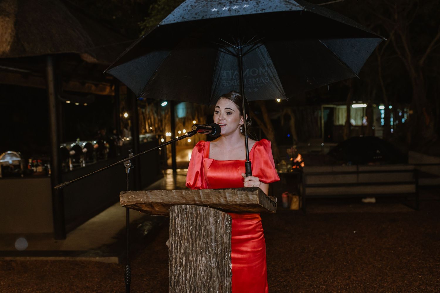 Woman in red satin dress speaks at wooden podium under black umbrella on rainy night street.