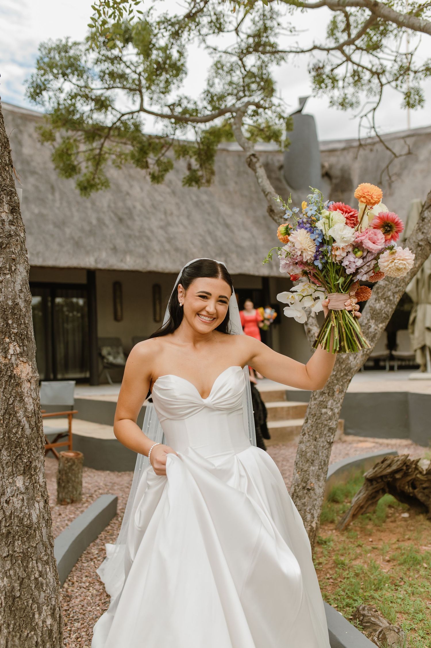 Smiling bride in strapless white ball gown holding a vibrant colorful bouquet beside a tree outdoors.
