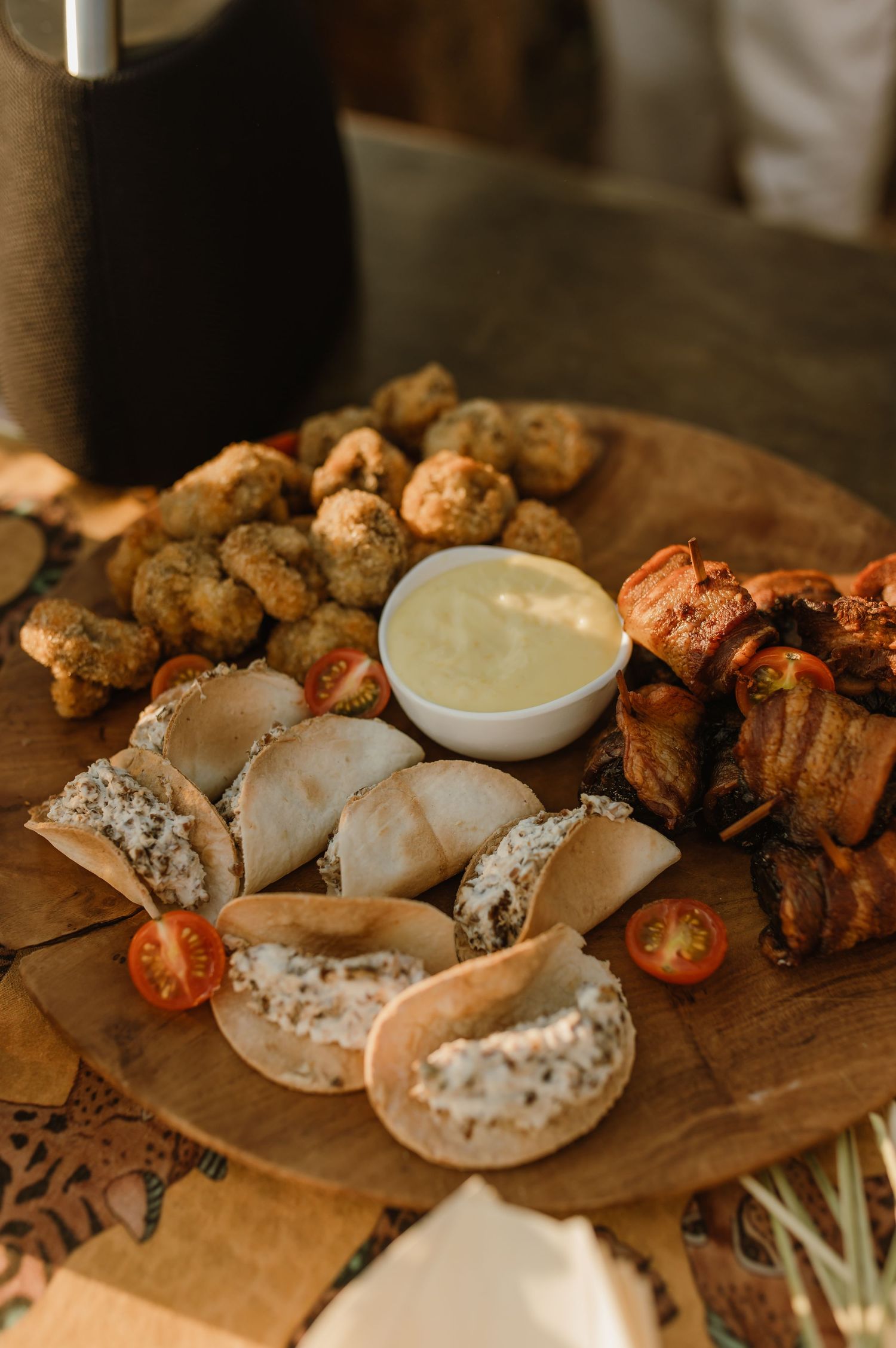 Rustic wooden board with fried appetizers, bacon-wrapped bites, pita chips, cherry tomatoes, and a creamy dipping sauce.