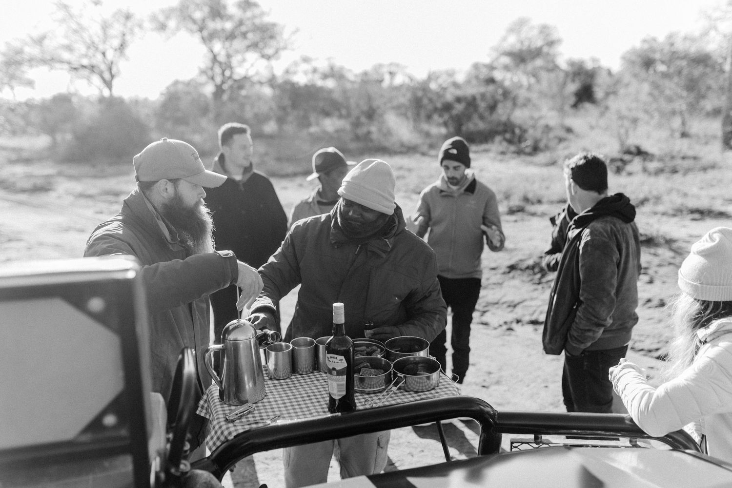 A group gathers around a safari vehicle in black and white photography during an outdoor expedition.