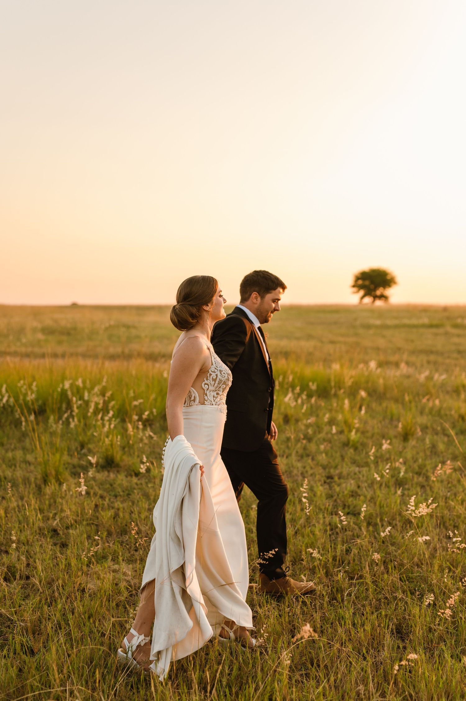 A couple walks through a golden field at sunset with a lone tree in the distance.