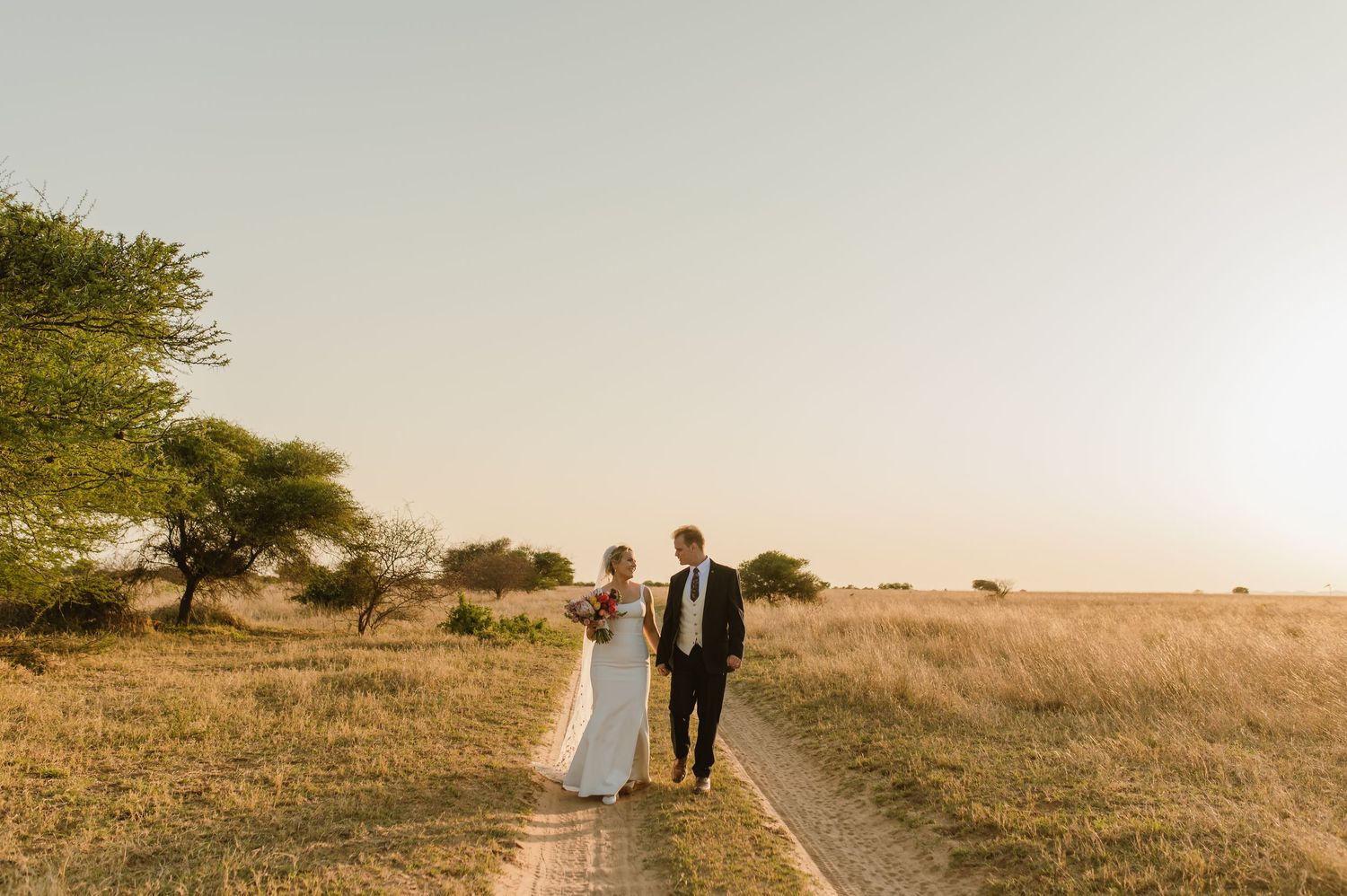 A romantic sunset wedding photoshoot in a golden field with a dirt path stretching into the distance.