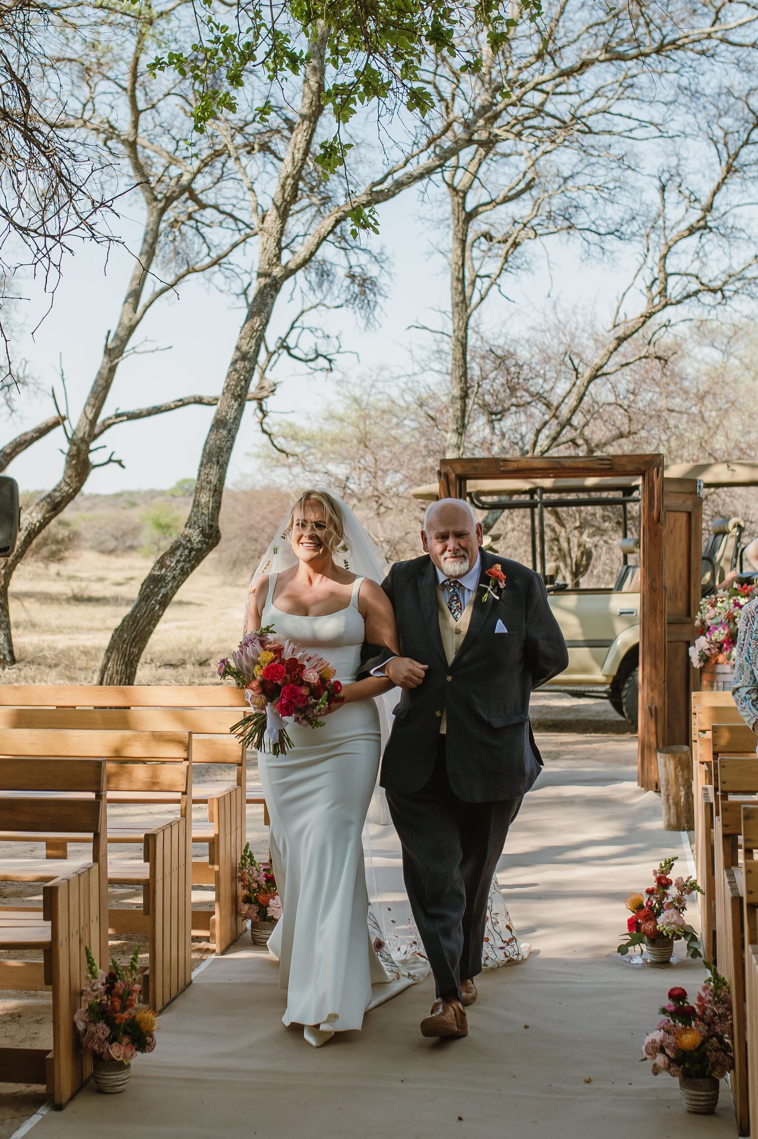 A couple laughs together on wooden benches during their rustic outdoor wedding ceremony.