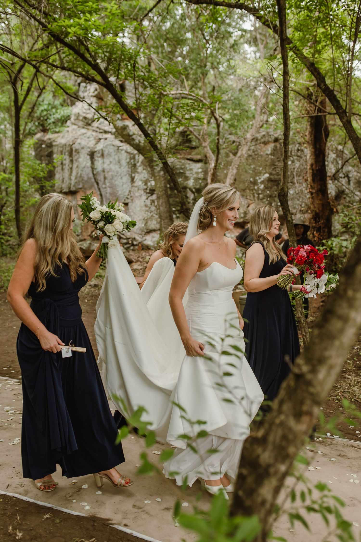 A bride in a white gown walks with her bridesmaids along a stone path surrounded by trees.
