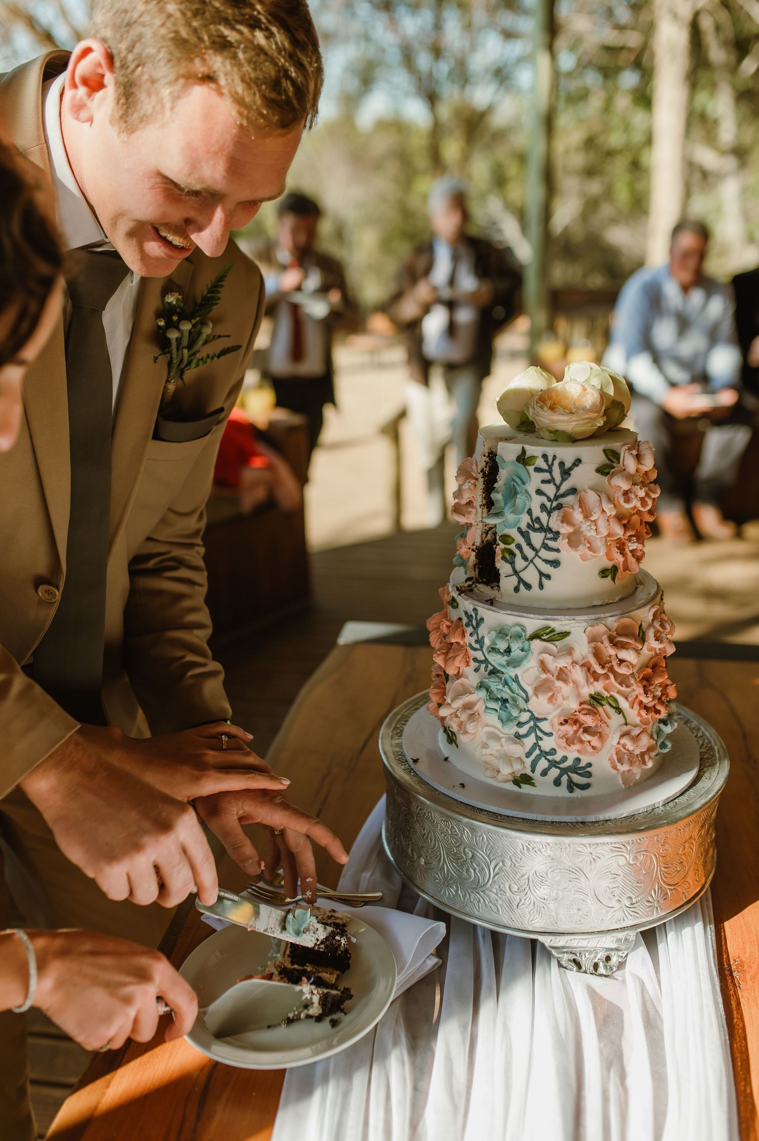 Wedding cake decorated with hand-painted flowers sits on silver cake stand during reception.
