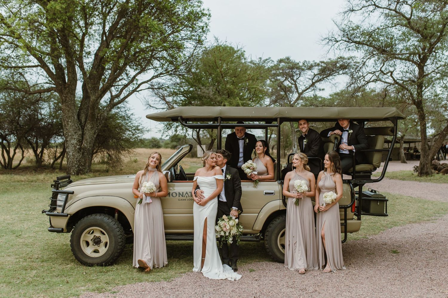 Wedding party poses near vintage vehicles in a rustic outdoor setting.