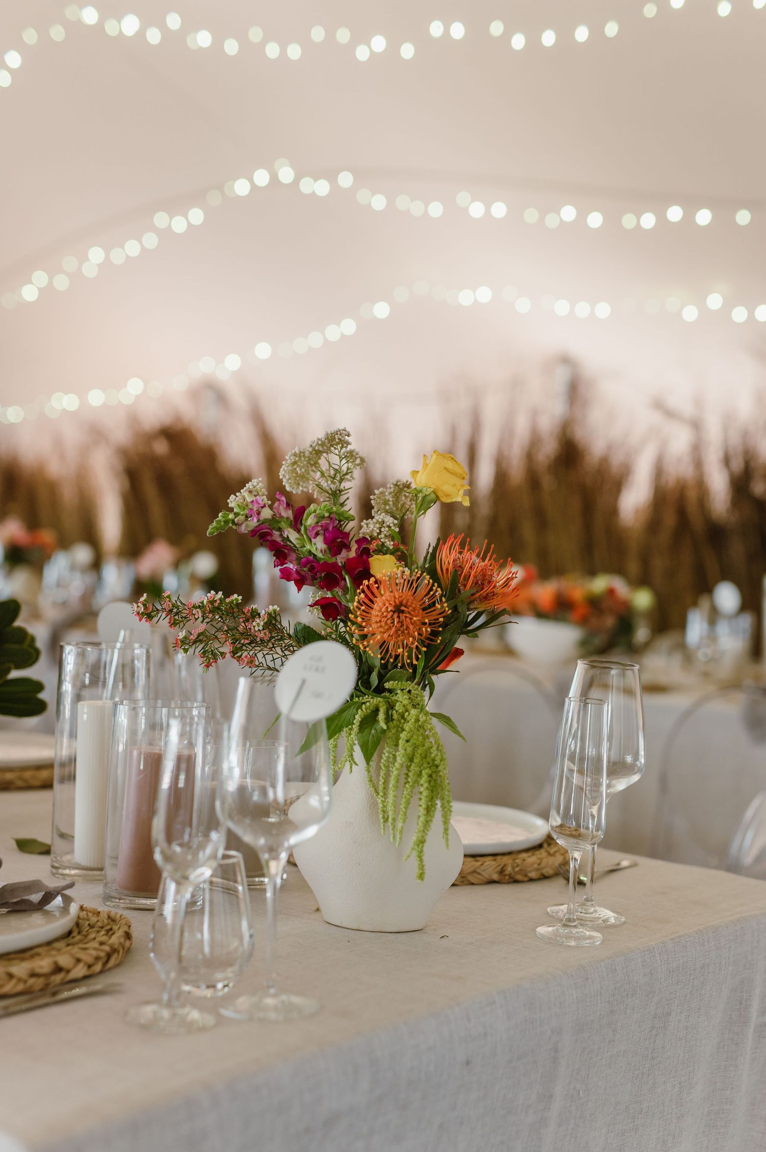 Wedding reception tables decorated with string lights, colorful florals, and glassware on linen tablecloths.