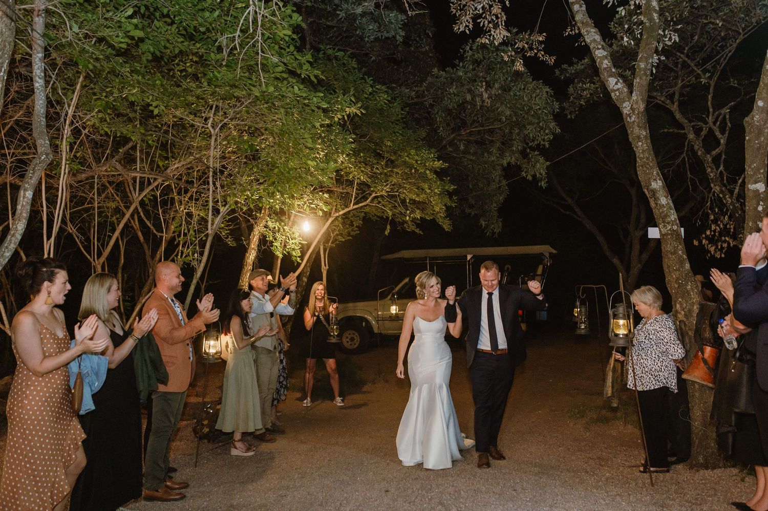 Outdoor celebration with guests gathered among trees and ambient lighting.