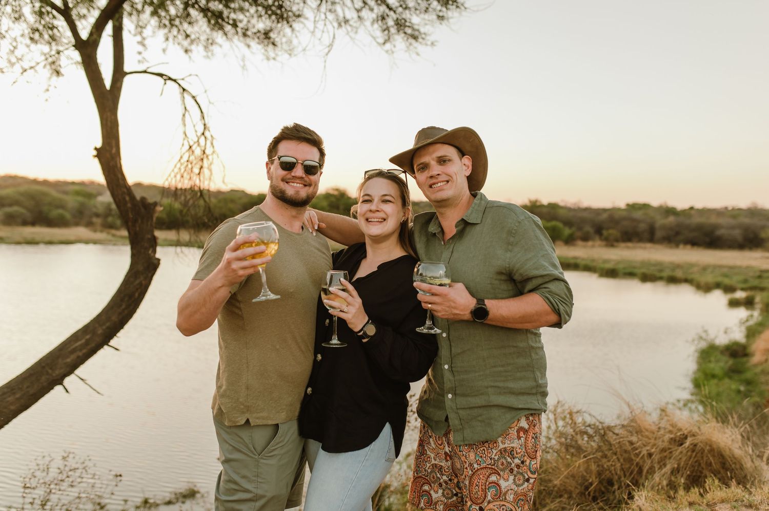 Group enjoying drinks together at sunset by a lake with trees in the background.