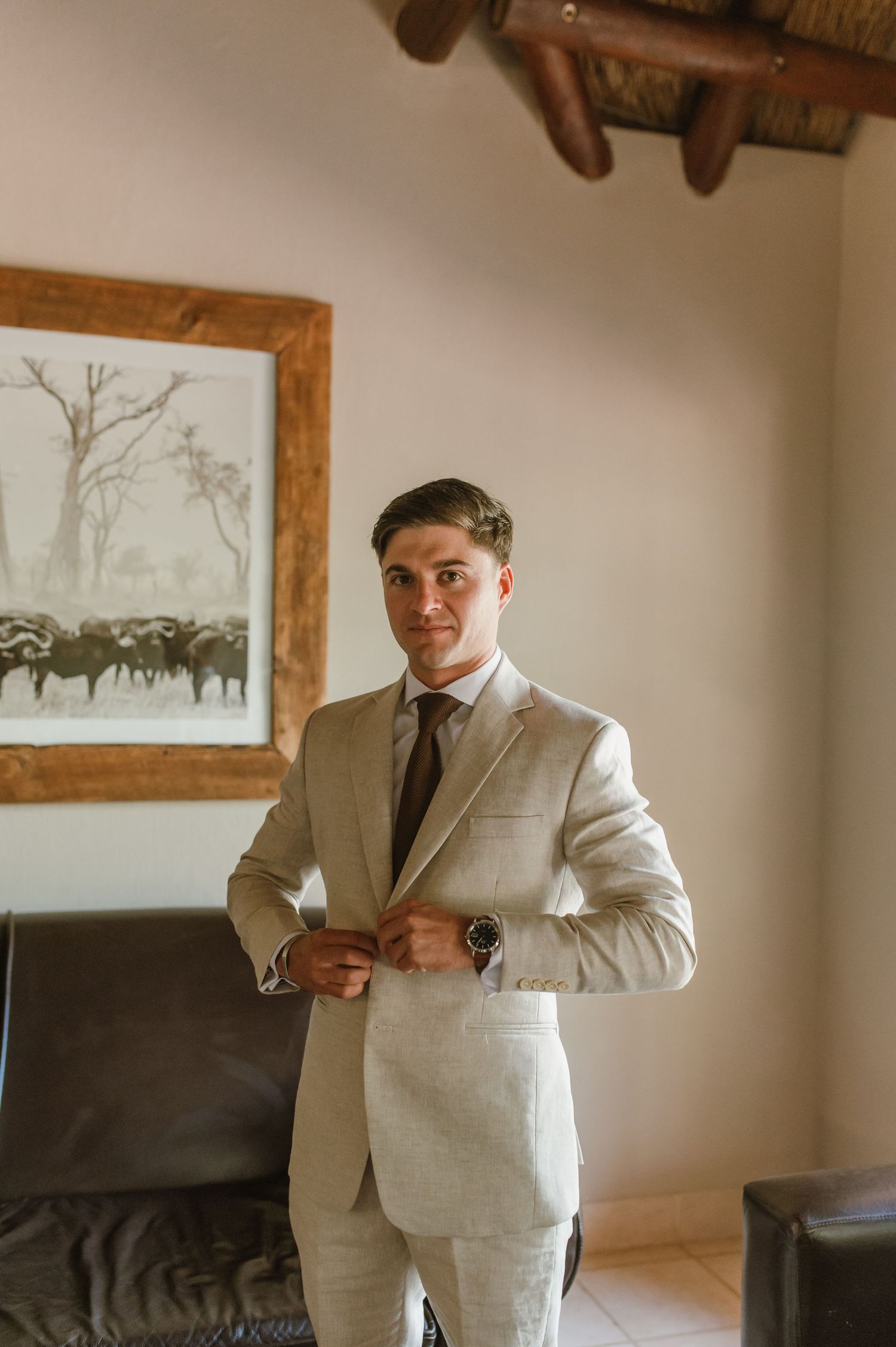 Someone in a light suit adjusting their tie in a room with rustic wooden beams.