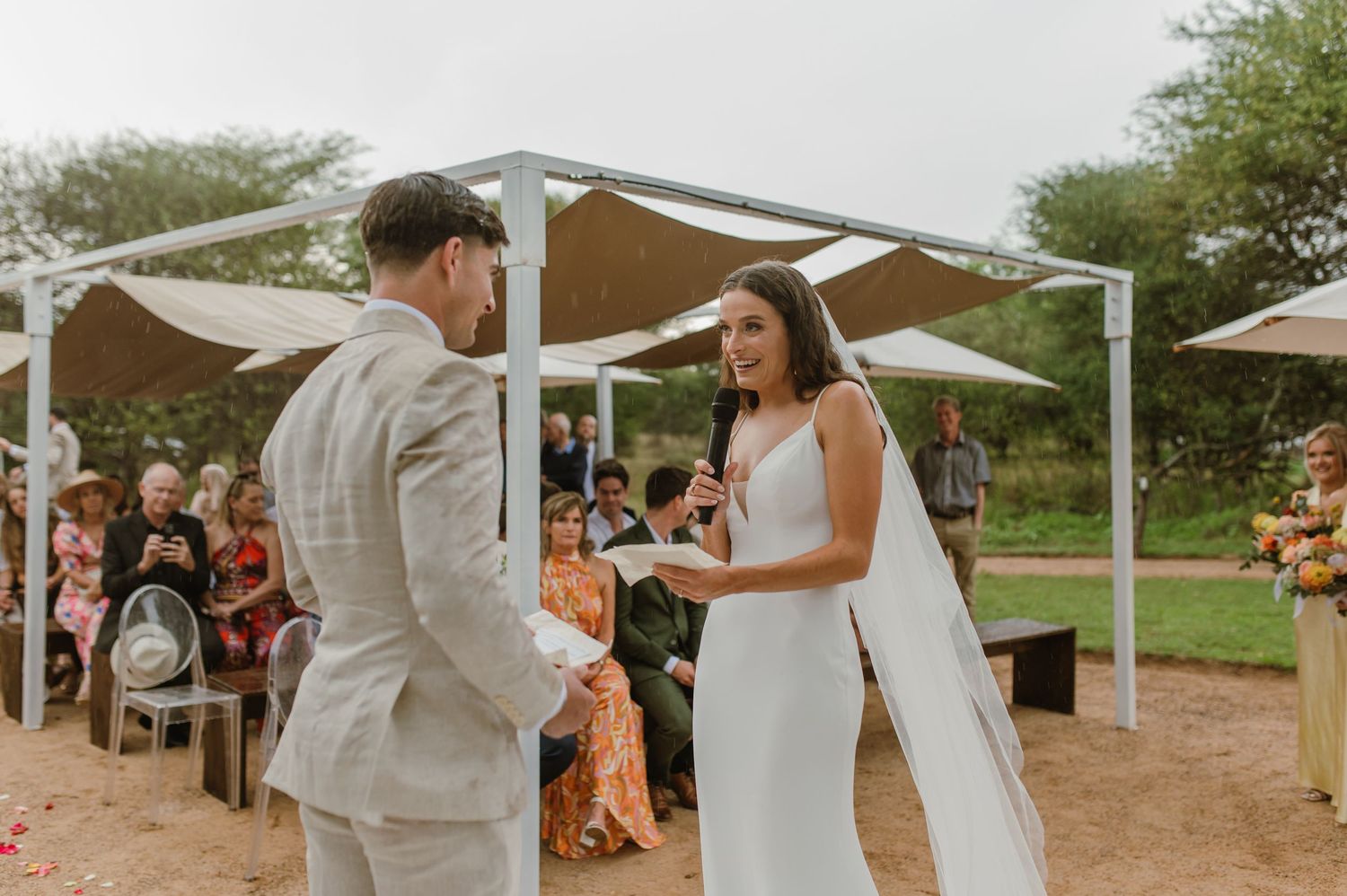 A wedding ceremony takes place under a white pergola with floral decorations in an outdoor setting.