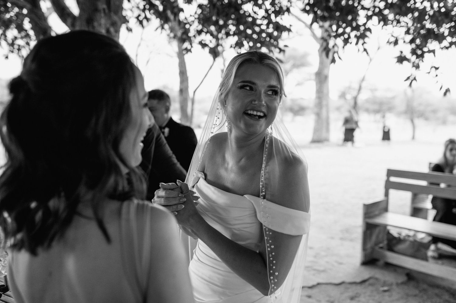 Joyful wedding celebration with guests seated on wooden benches under shady trees.