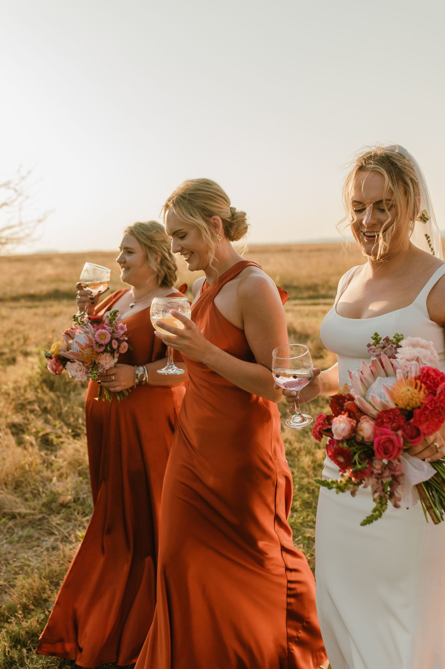 Wedding party enjoys drinks and laughter while holding colorful floral bouquets at sunset.