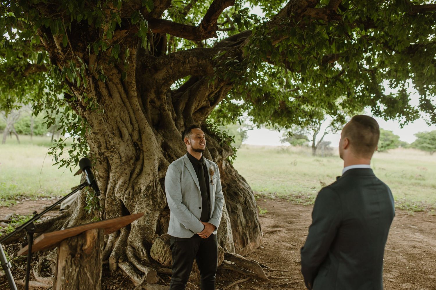 Two men having a conversation under a large tree at an outdoor wedding venue.