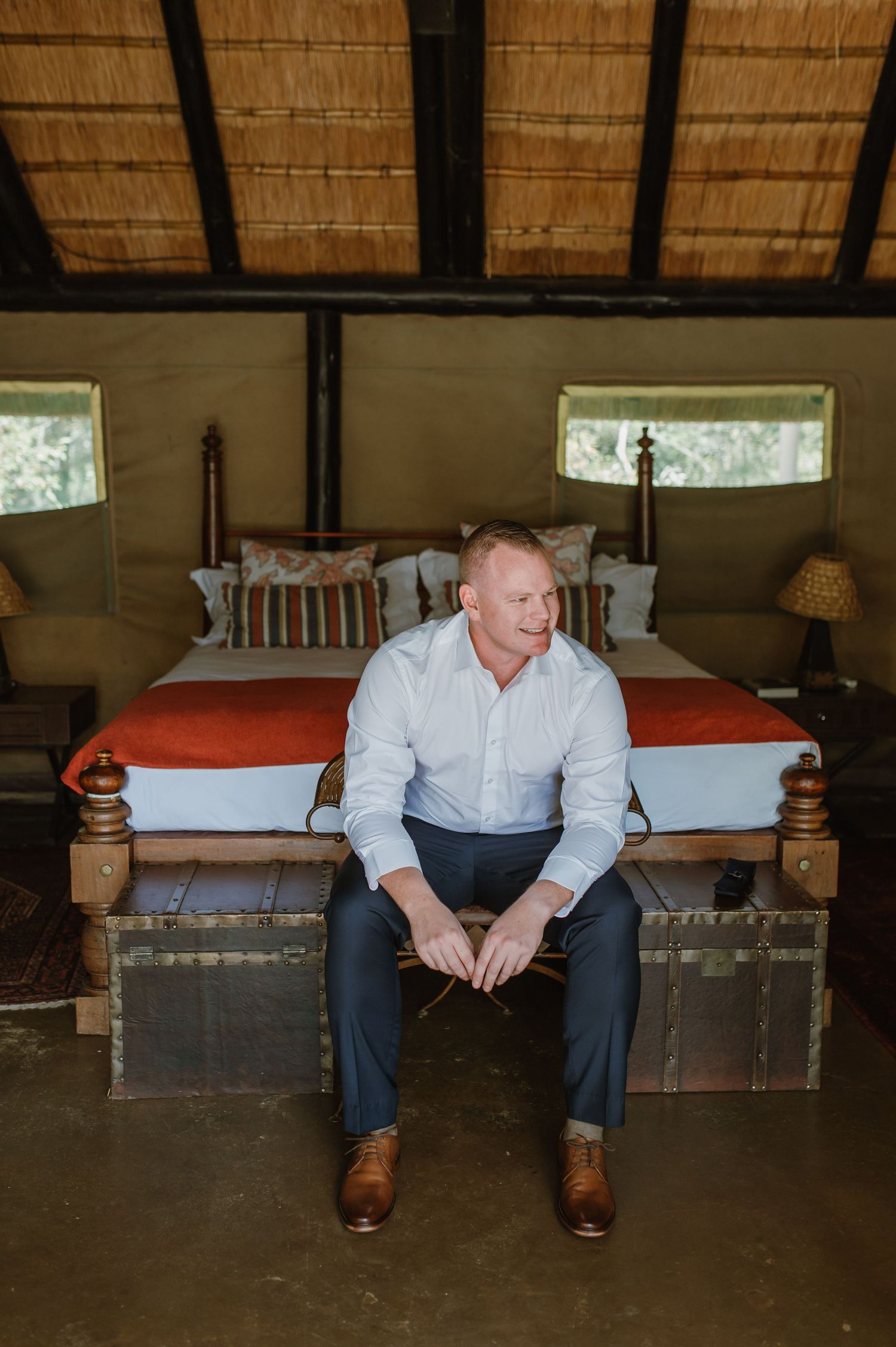 Person in white shirt and dark pants sitting on vintage trunk under thatched roof.