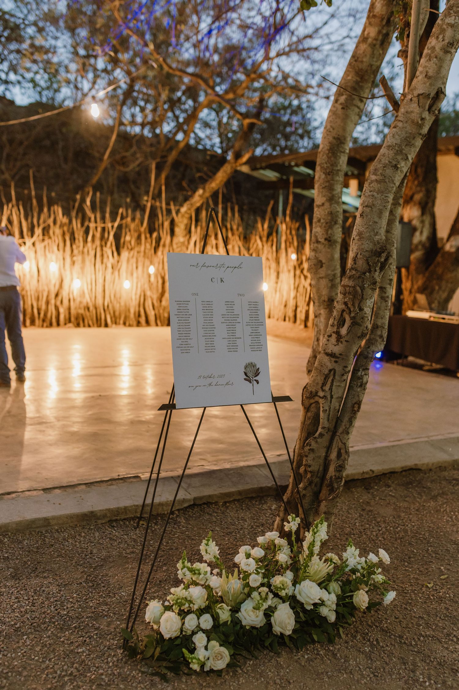 Wedding seating chart display with white floral arrangements and outdoor lighting at dusk.