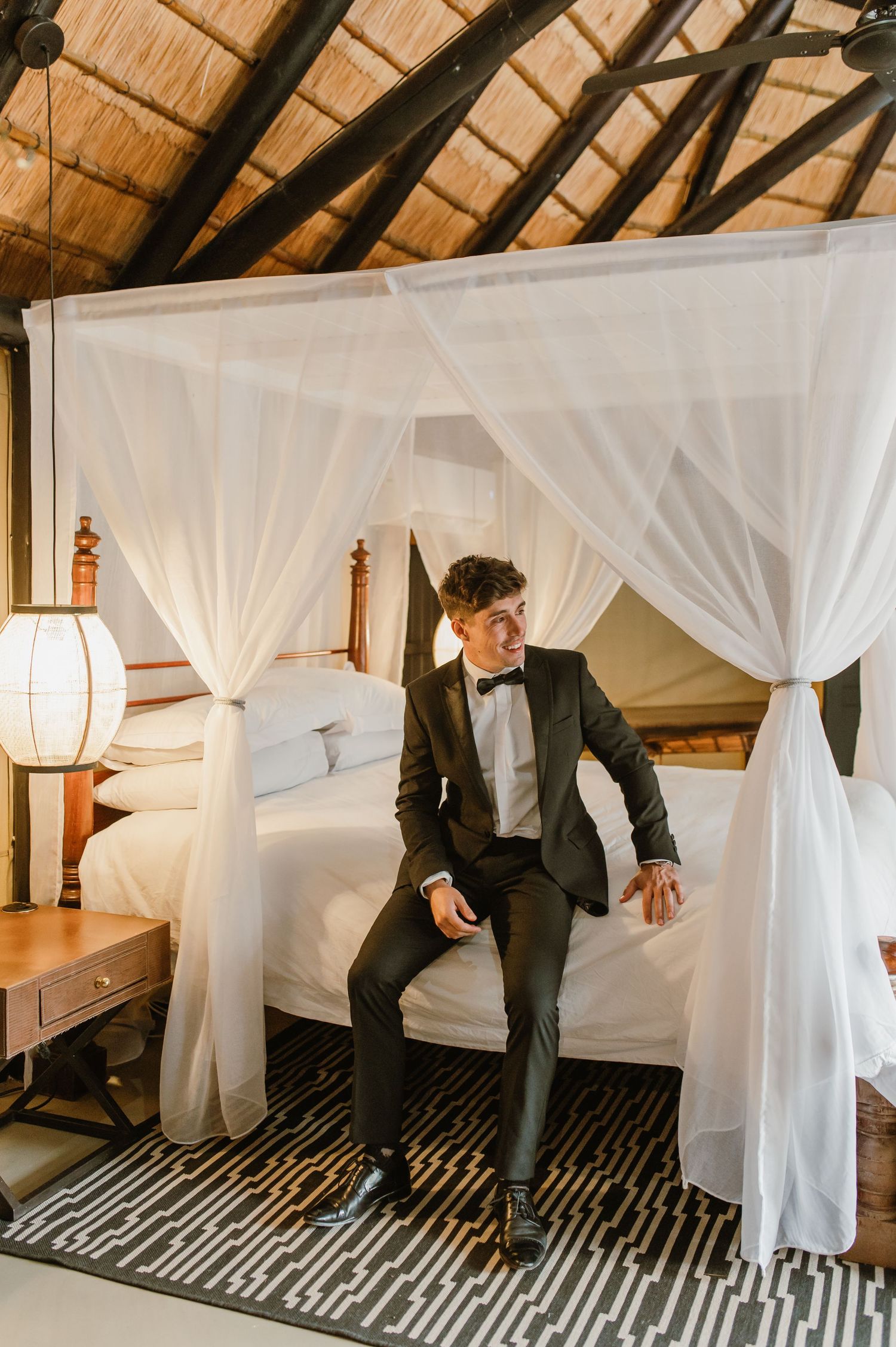 Elegant scene with someone seated on a canopy bed with white drapes and wooden beams overhead.