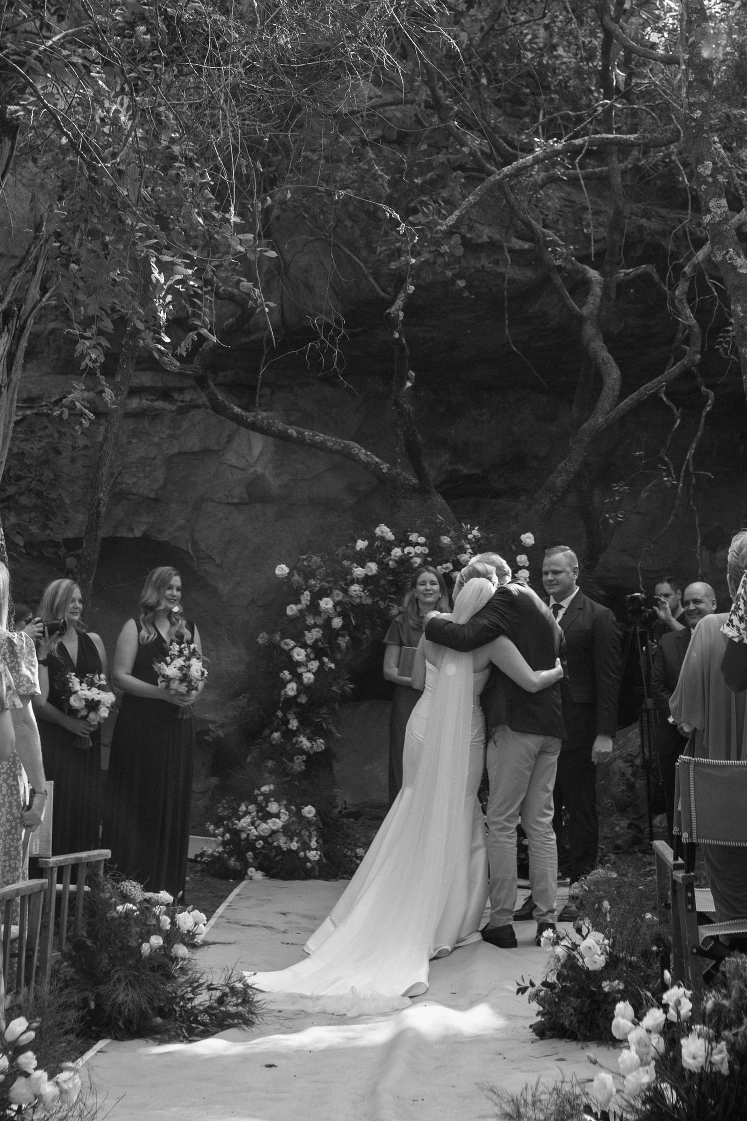 Black and white photo of a wedding ceremony under a floral arch in a forest setting.