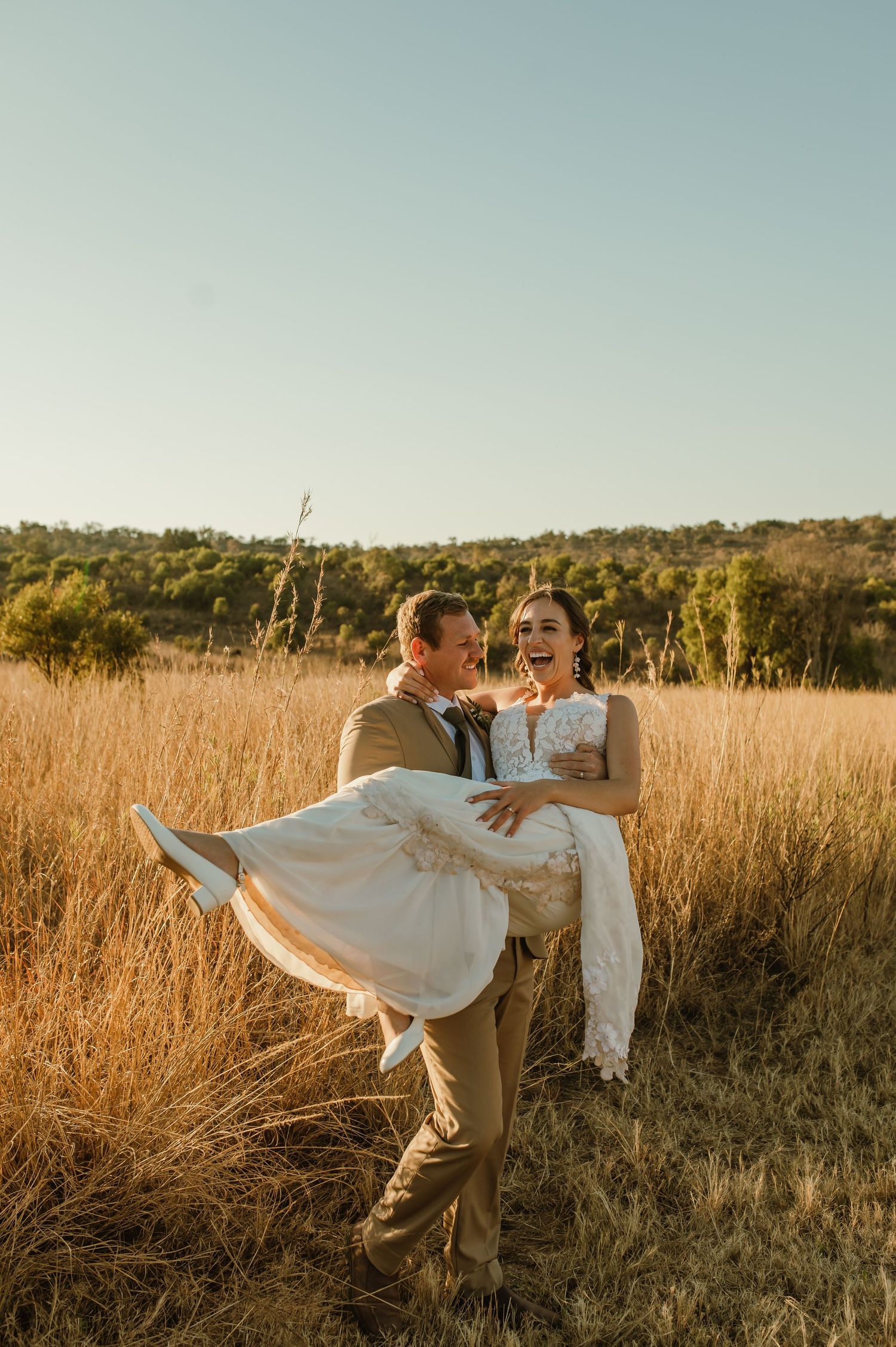 Groom carries bride in flowing white dress through golden field at sunset.