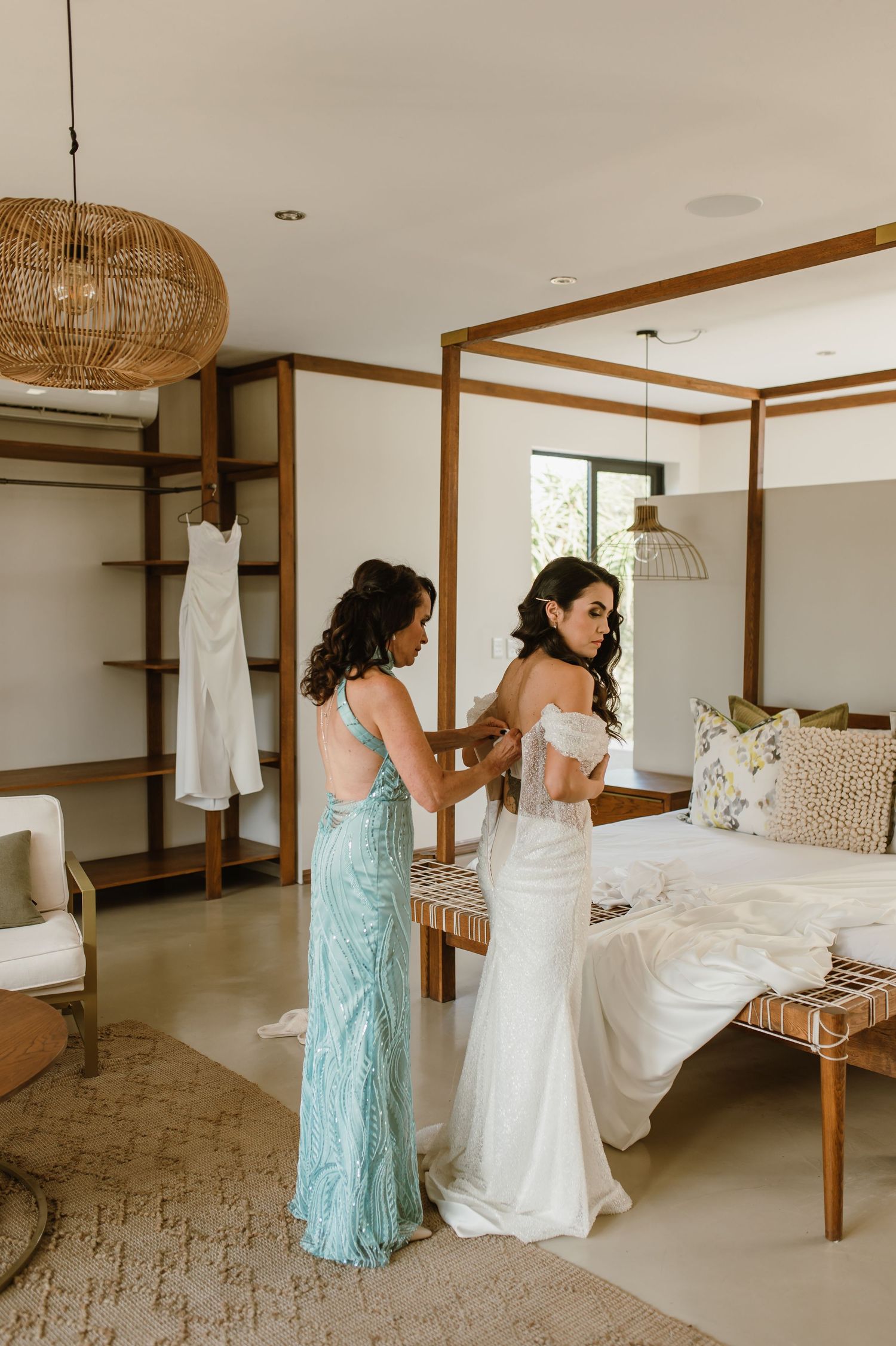 Two people helping with a white dress in a modern bedroom with woven pendant lights.