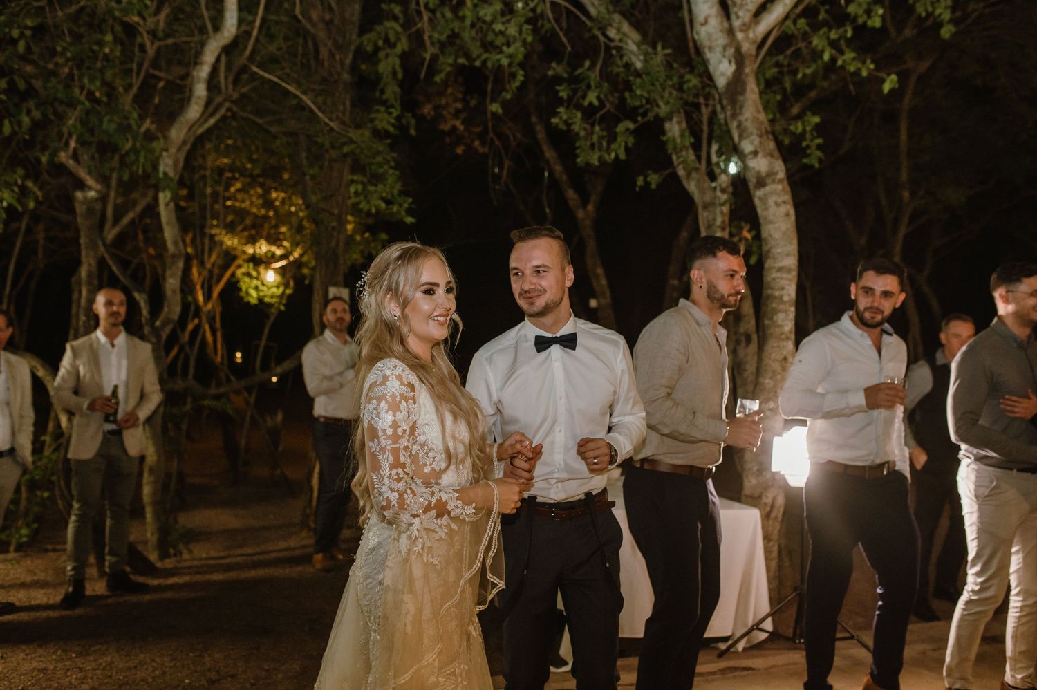 Wedding celebration at night with guests dancing under trees and ambient lighting.