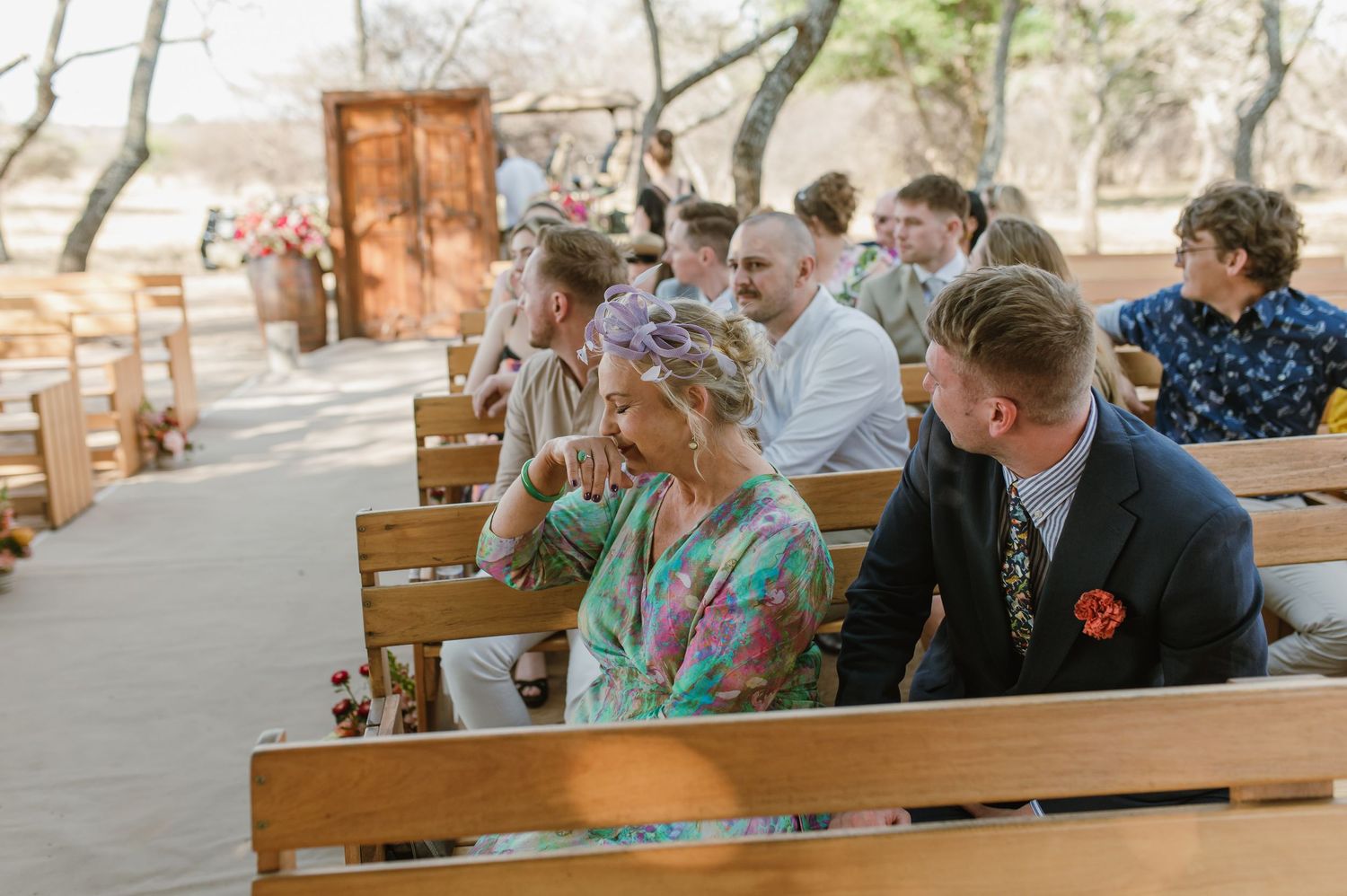 Wedding guests seated on wooden benches smile and watch the ceremony unfold.