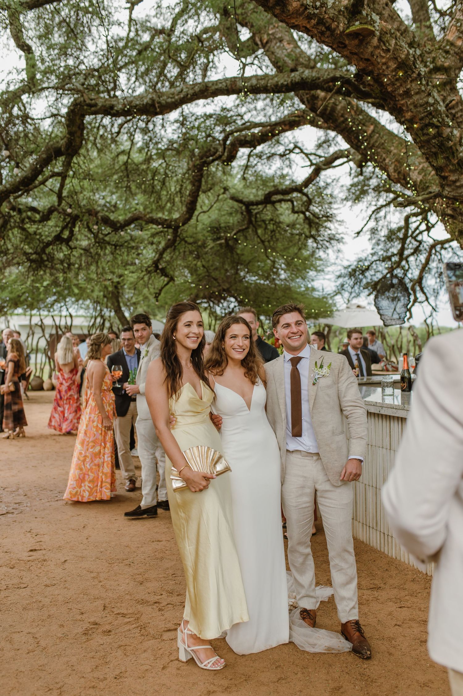 Wedding party and guests in light summer attire pose for photos under shaded trees.