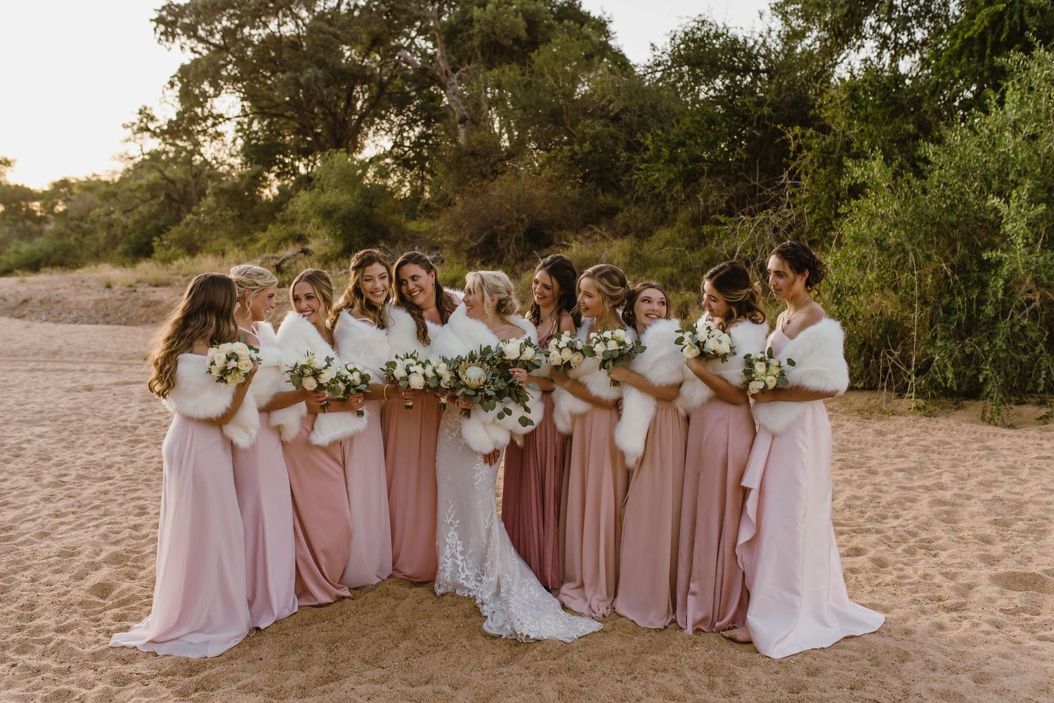 Bridesmaids in flowing pink dresses gather for group photo on sandy beach.