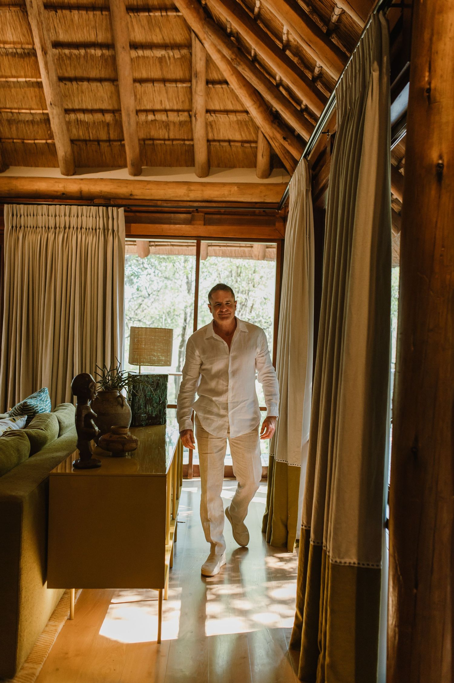 Man in white linen outfit walking through a luxury safari lodge with thatched roof and wooden interiors.