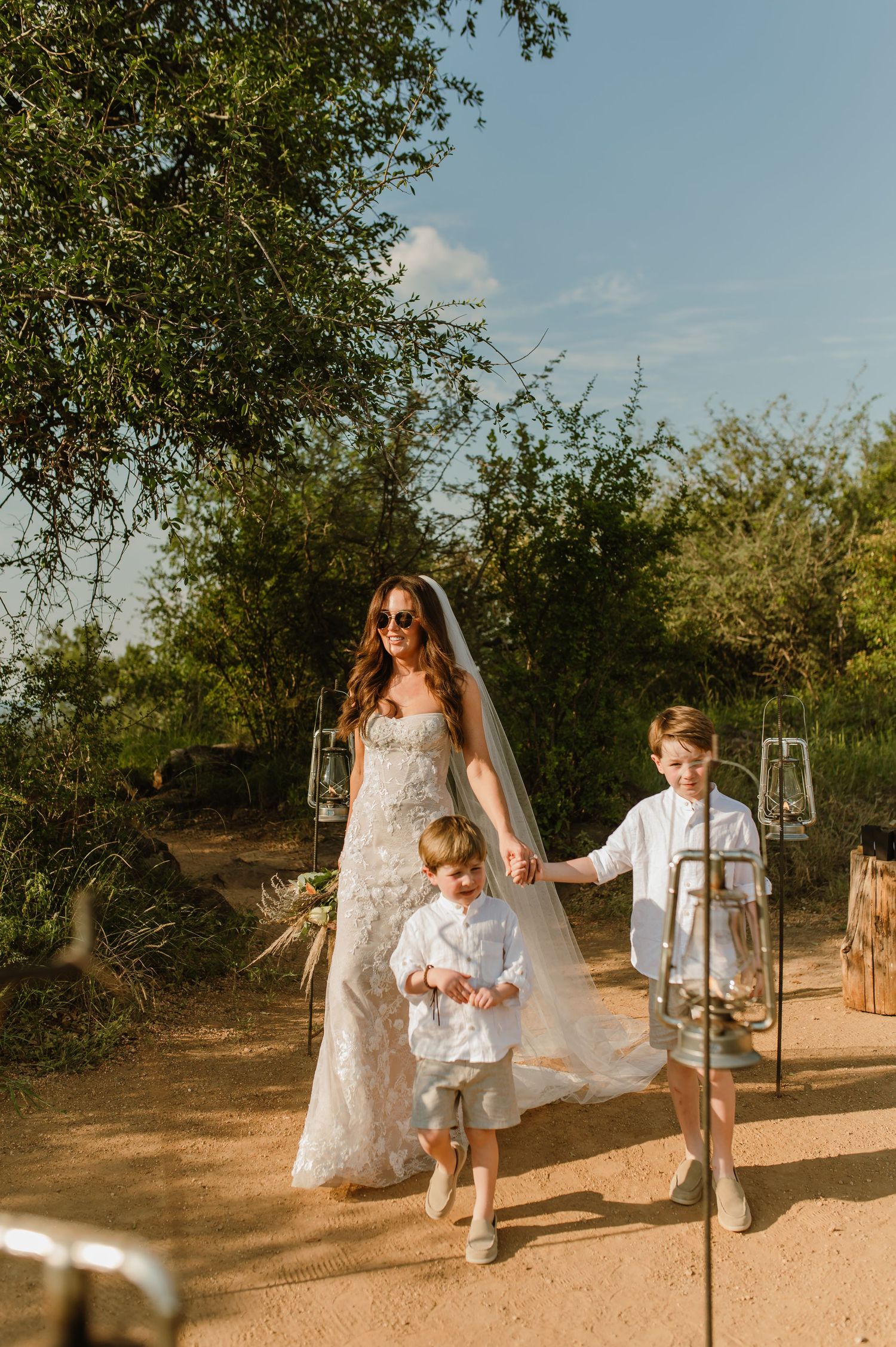 Bride in lace wedding dress and veil walking with two young boys as page boys outdoors.