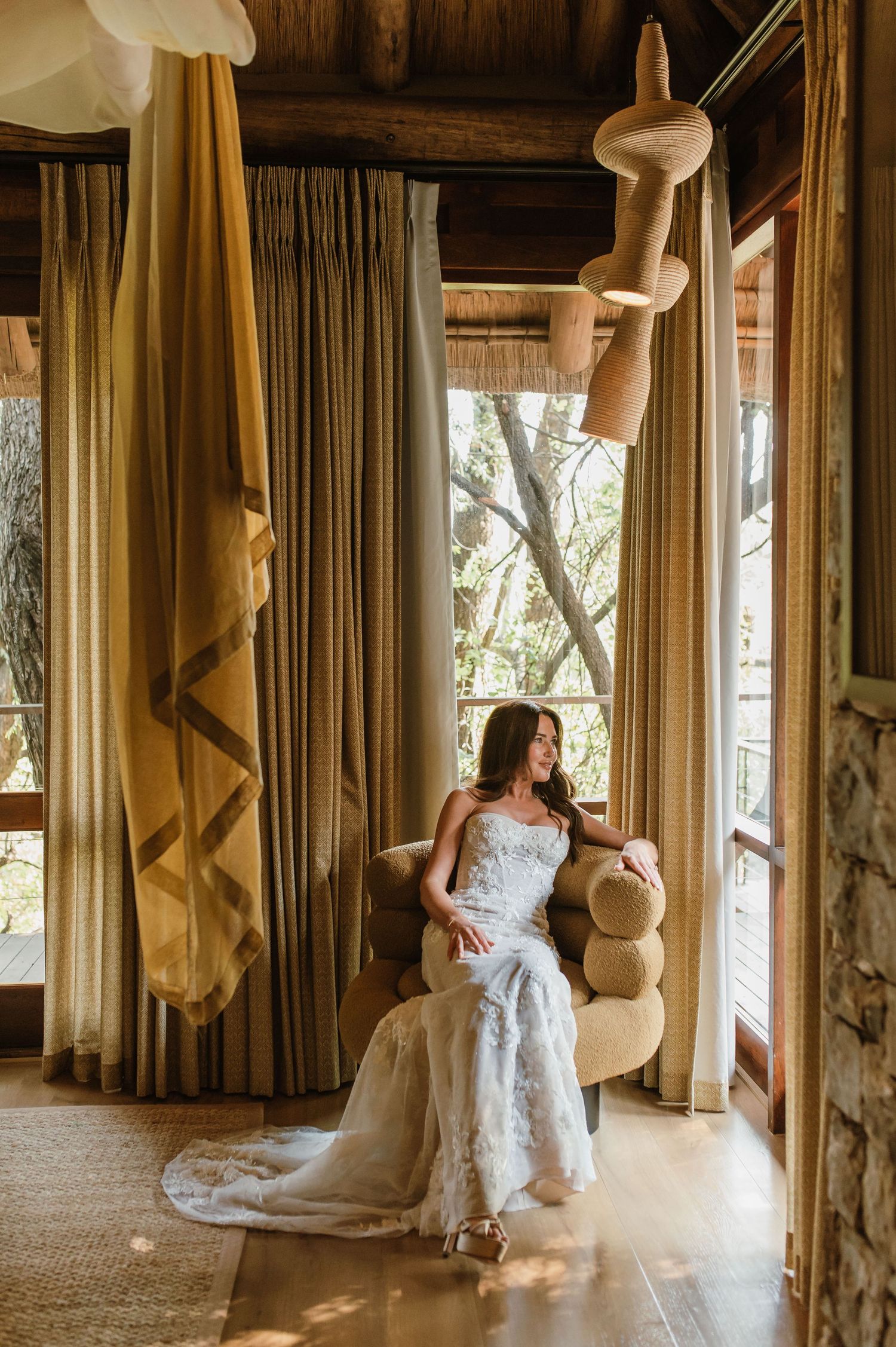 Elegant bride in a lace wedding gown seated in a chair by large windows in a luxury safari lodge.