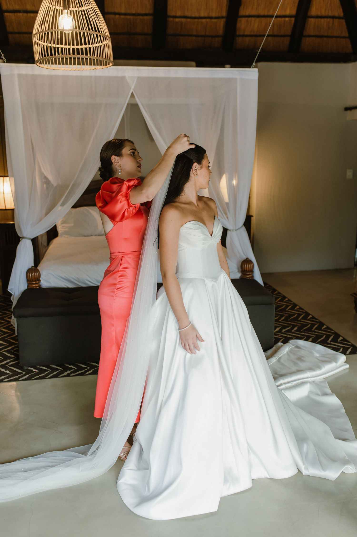 Bridesmaid in coral dress adjusting bride's veil near a canopy bed in a safari lodge bridal suite.
