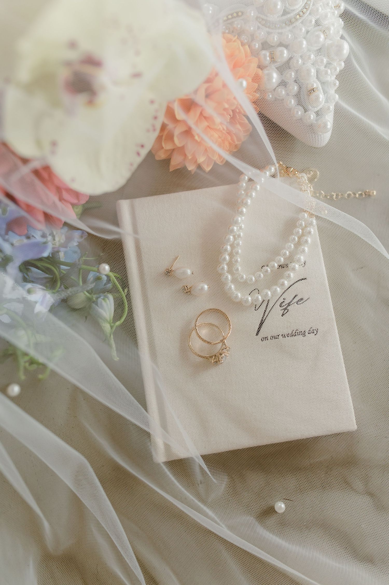 Elegant bridal flat lay with pearl necklace, gold rings, earrings, and a Wife wedding day book on ivory fabric.