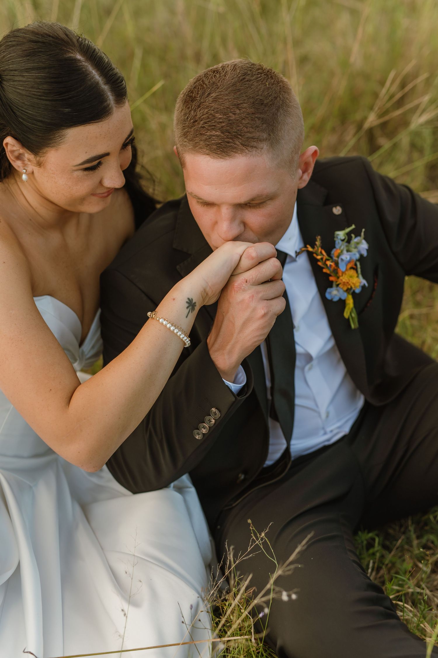 Groom in black suit kisses bride's hand while sitting in grass during romantic outdoor wedding portrait.