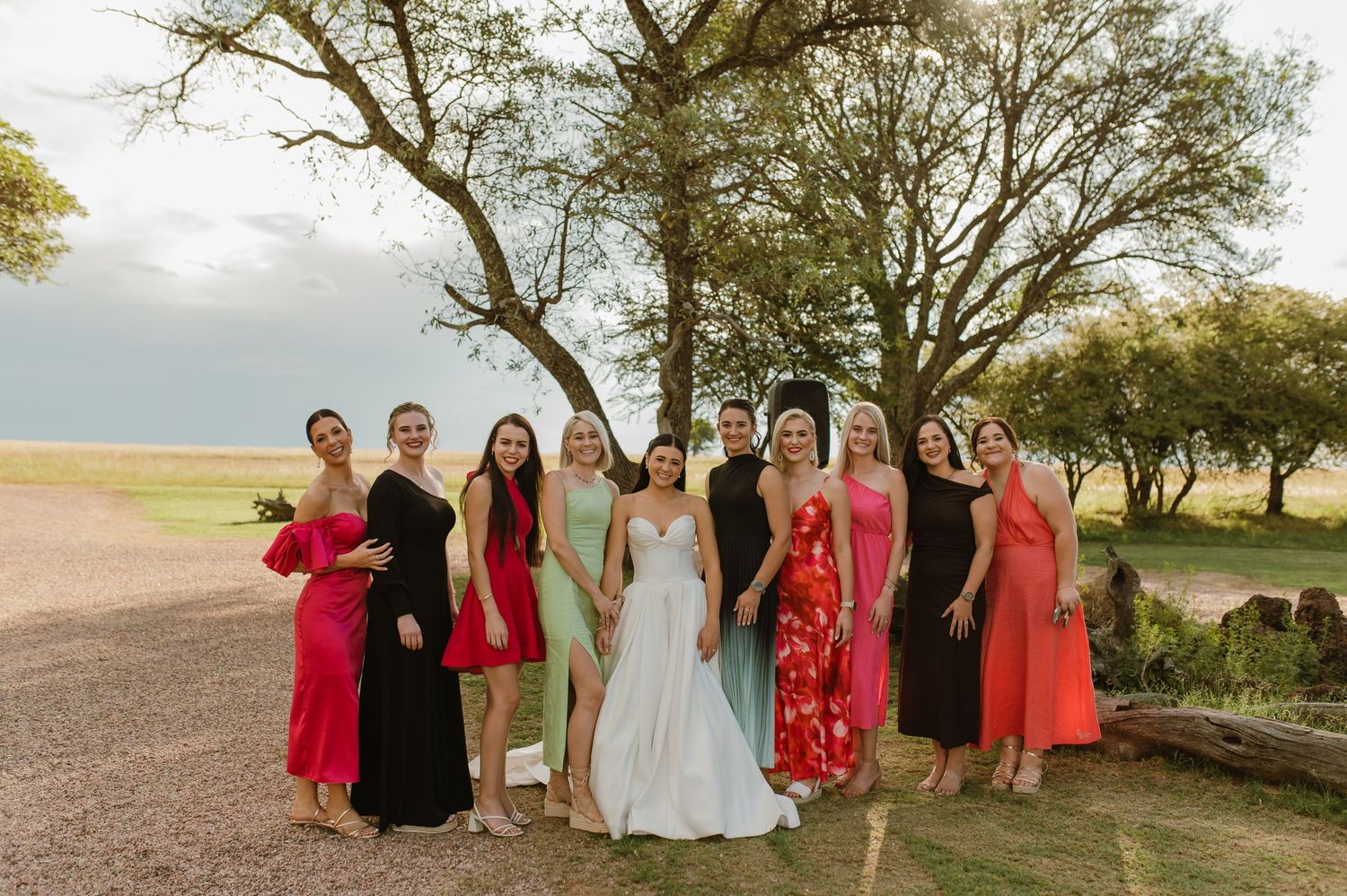 Bride in white gown poses with wedding guests in colorful dresses outdoors near trees on her wedding day.