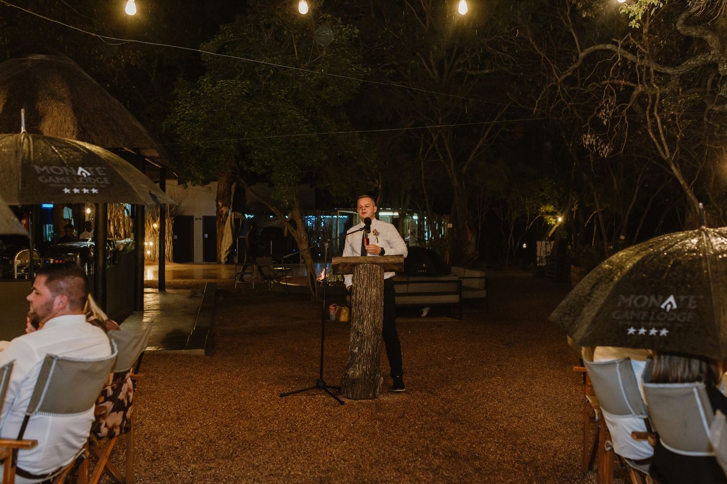 Wedding officiant speaks at podium microphone during outdoor evening ceremony under trees with string lights.