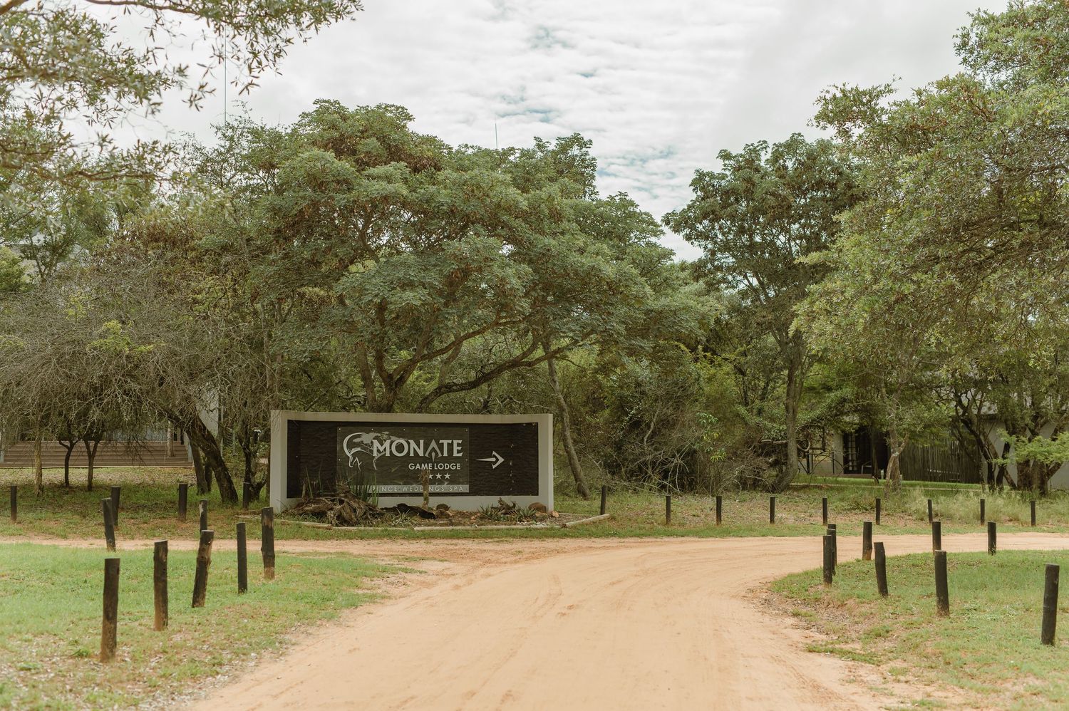 Entrance gate to Monate Game Lodge with a dirt road lined by wooden posts and lush trees under a cloudy sky.