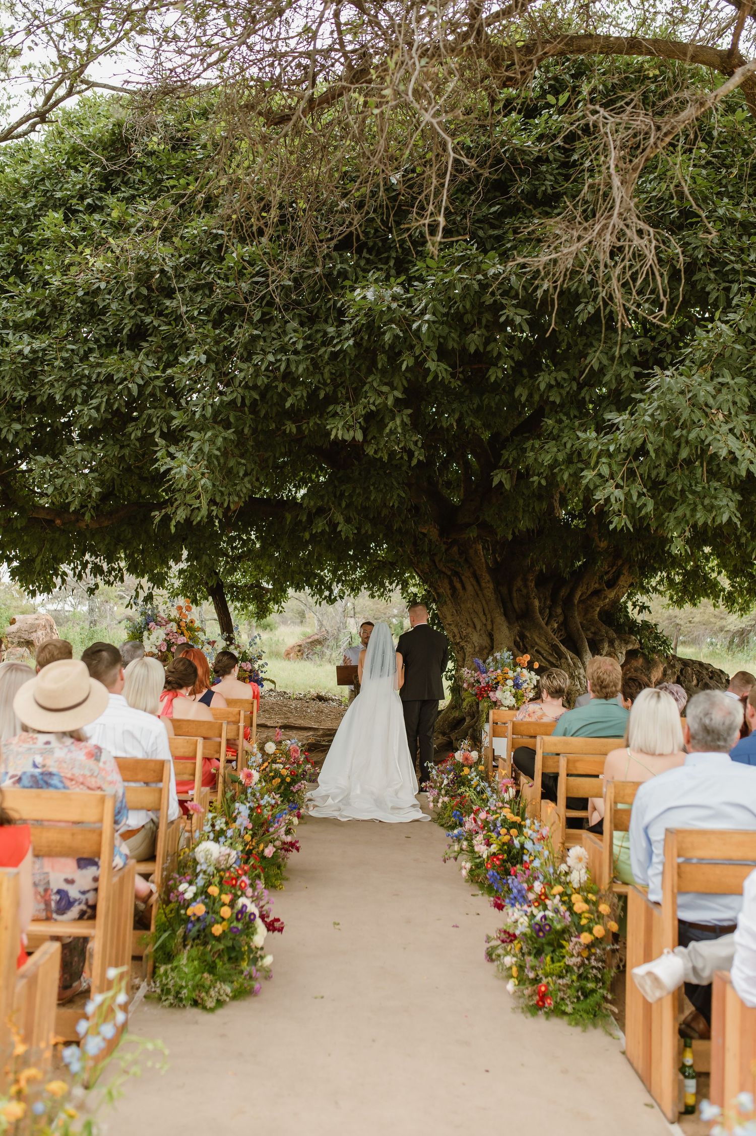 Outdoor wedding ceremony aisle lined with colorful wildflowers beneath a large tree.
