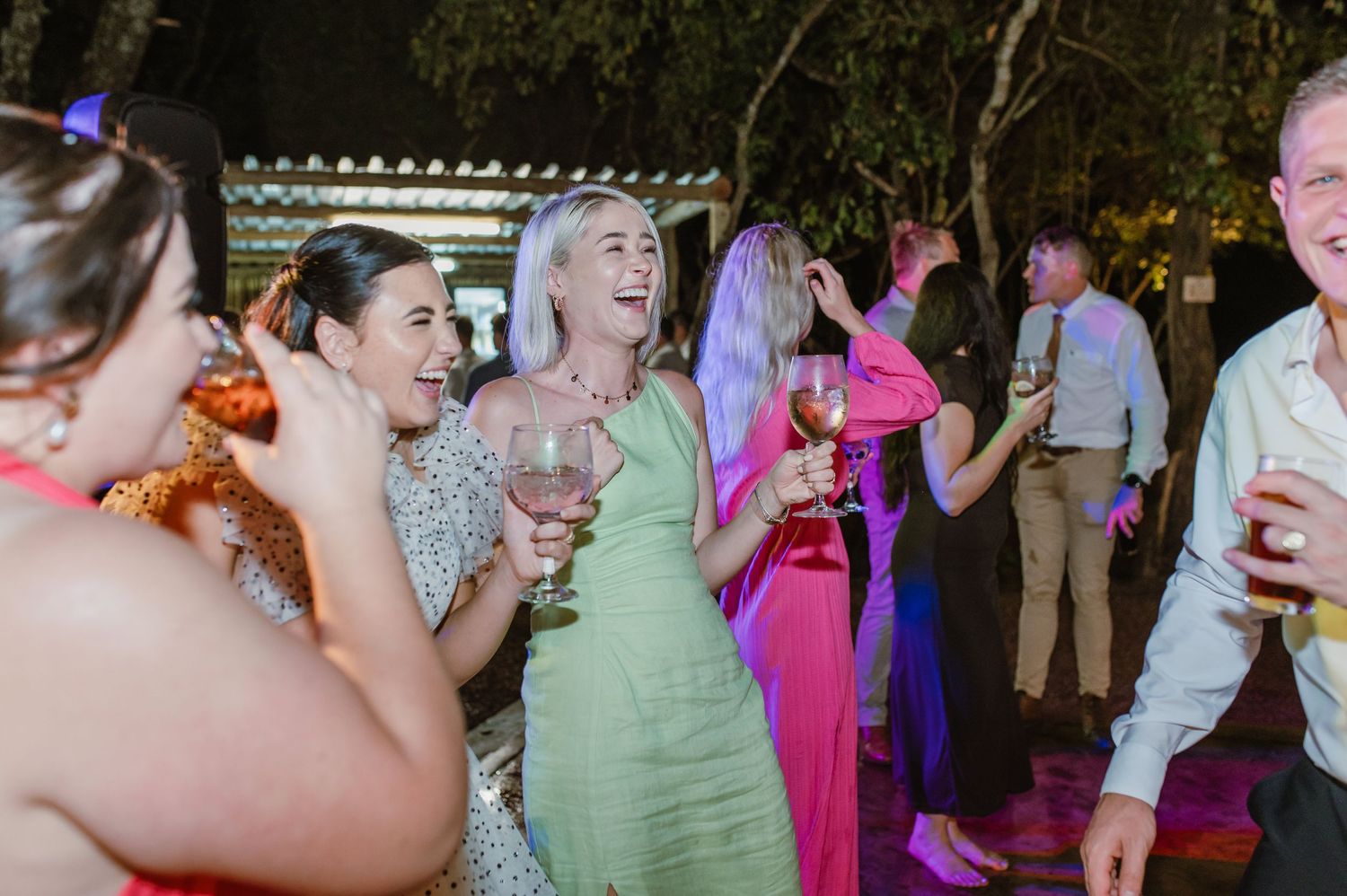 Happy wedding guests laughing and holding wine glasses while dancing at outdoor evening reception.