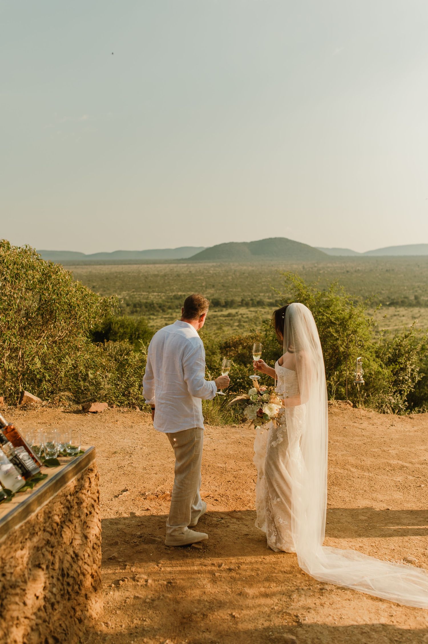Bride and groom sharing champagne toast at outdoor safari wedding with scenic African landscape and mountains in background.