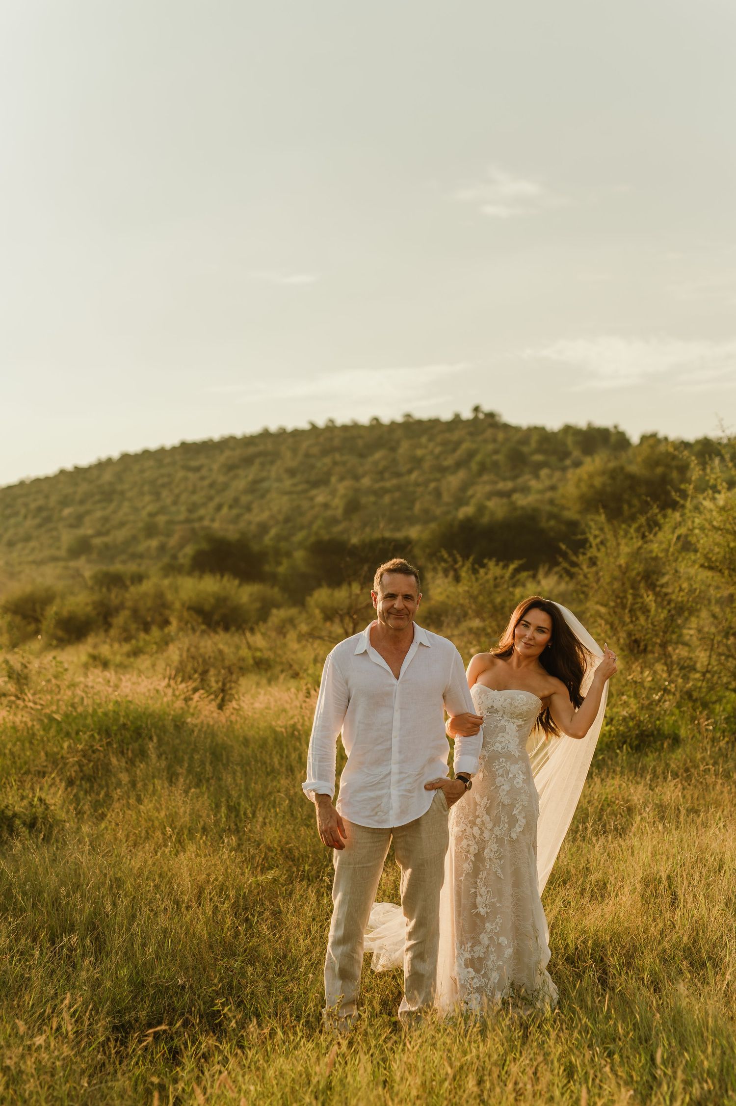 Bride and groom standing in golden sunlit field with green hills backdrop during outdoor wedding.