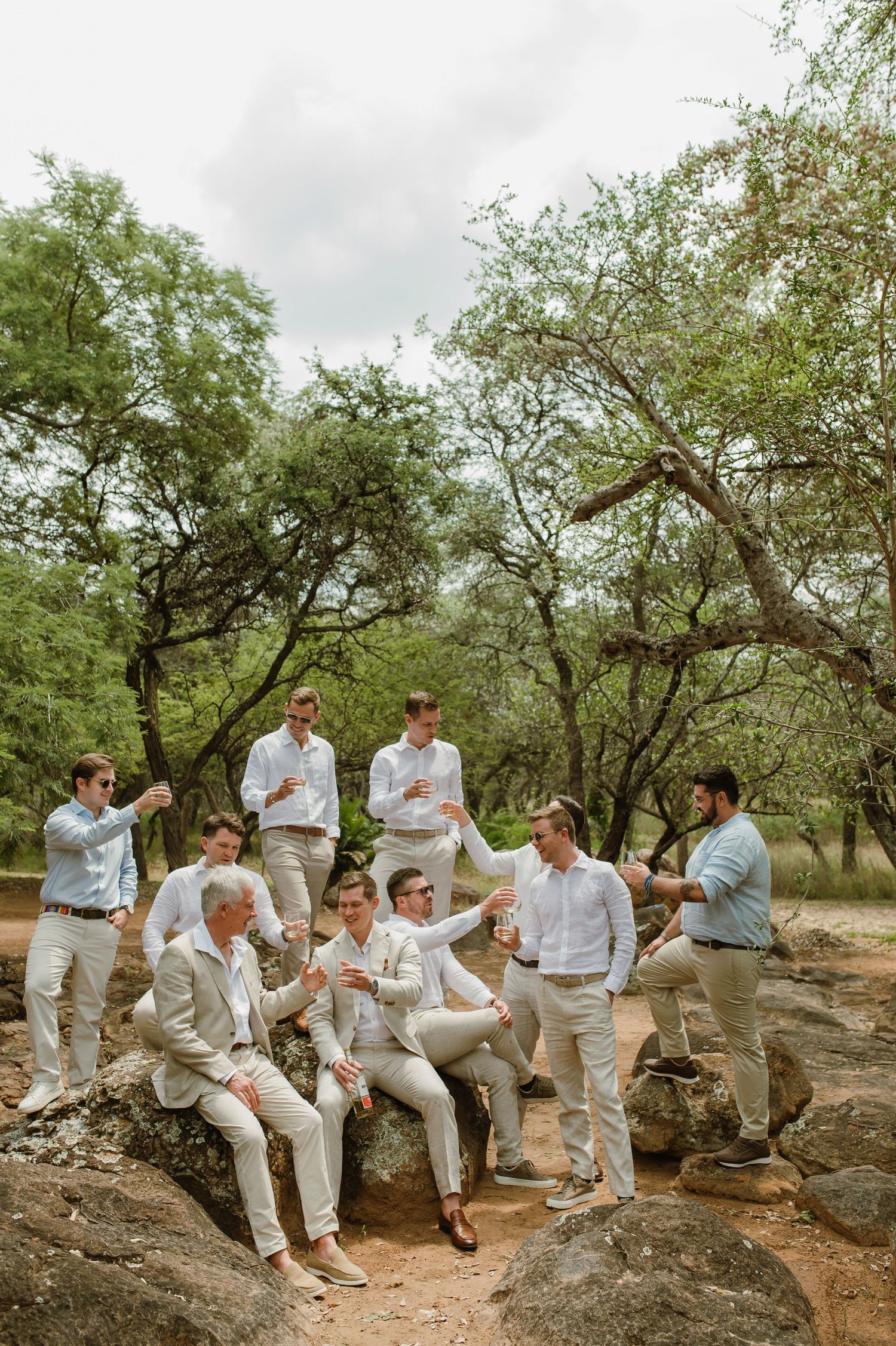 A group in light colored casual attire gathers on rocky terrain surrounded by trees on an overcast day.