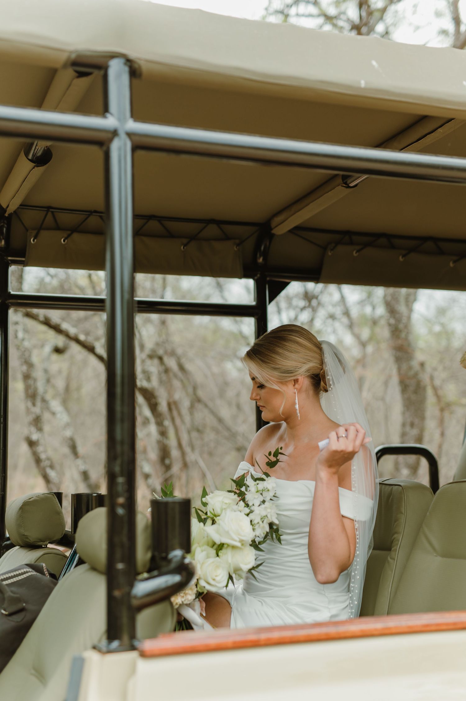 A bride in a white dress holding a bouquet sits inside a safari vehicle on her wedding day.