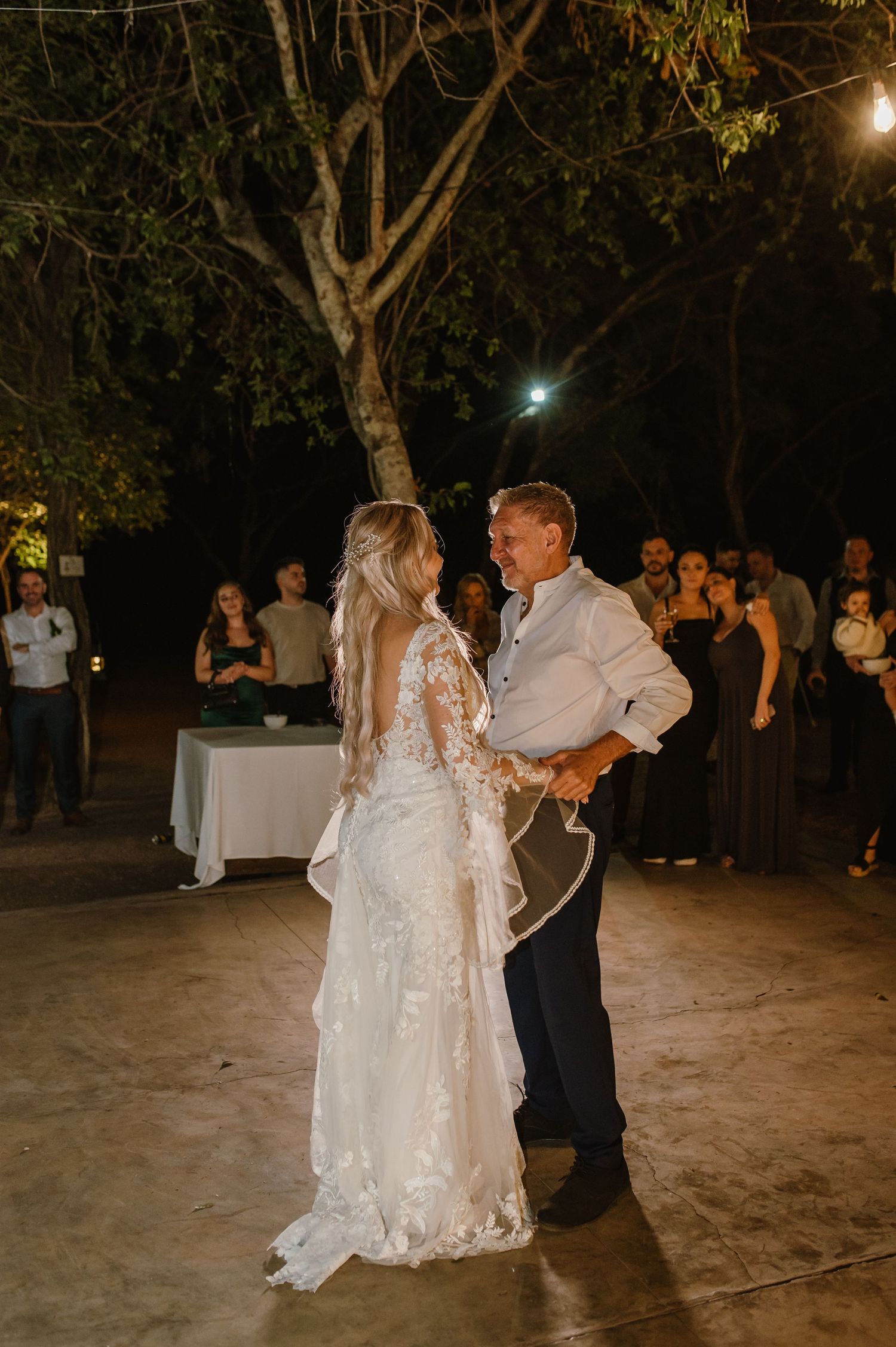 Couple sharing first dance on outdoor patio under trees and string lights at evening reception.
