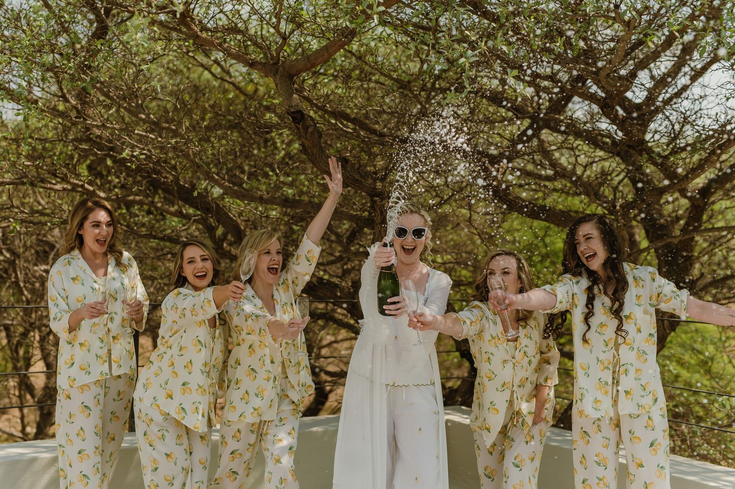 Group photo of bridesmaids in floral robes posing under blooming trees.
