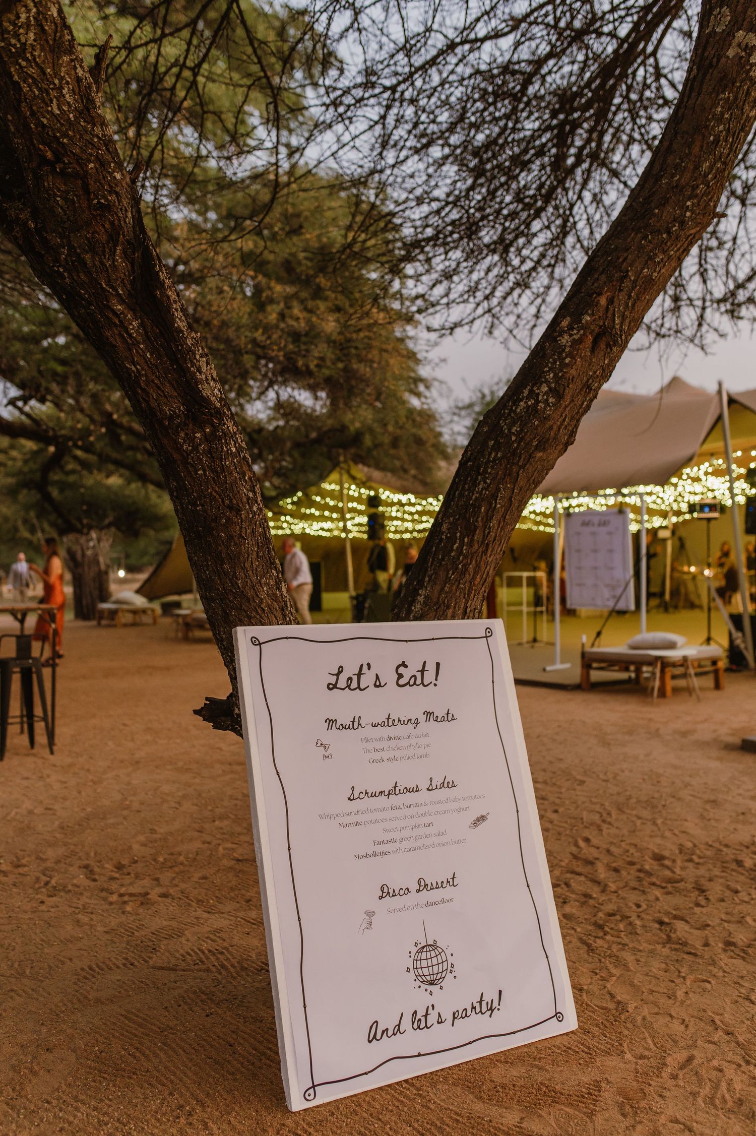 Wedding reception signage displayed under trees with twinkling lights at dusk.