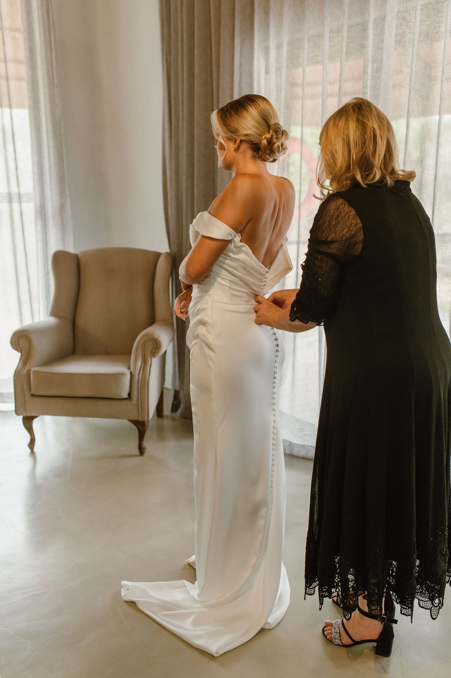 People in formal attire helping with dress preparations in bright indoor setting.