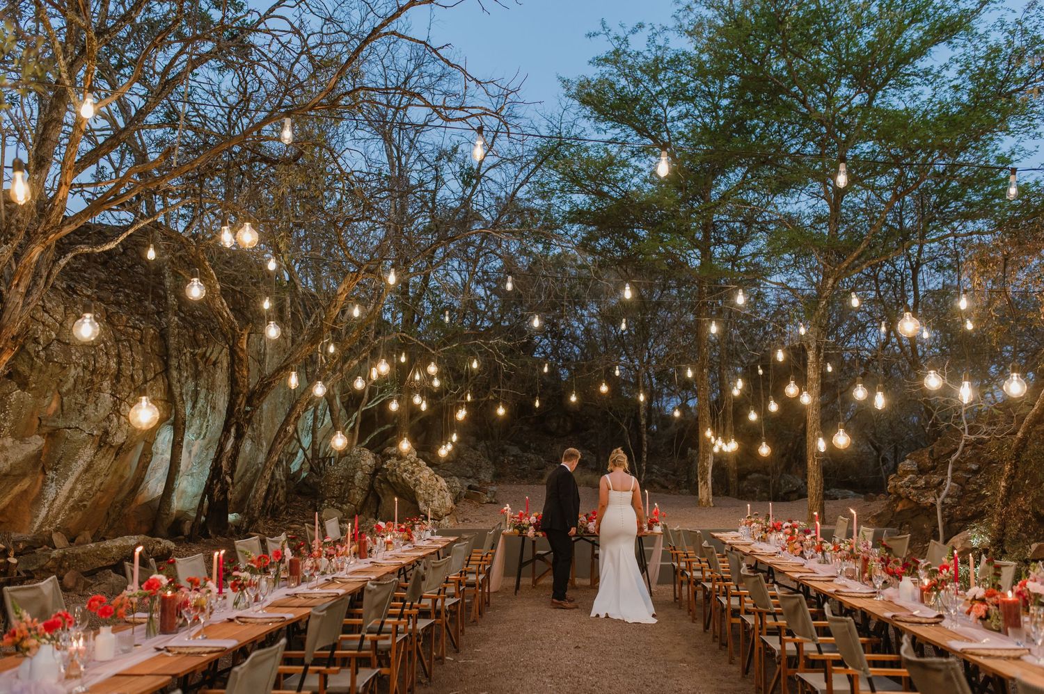 Long tables set for an outdoor wedding reception under string lights and trees.