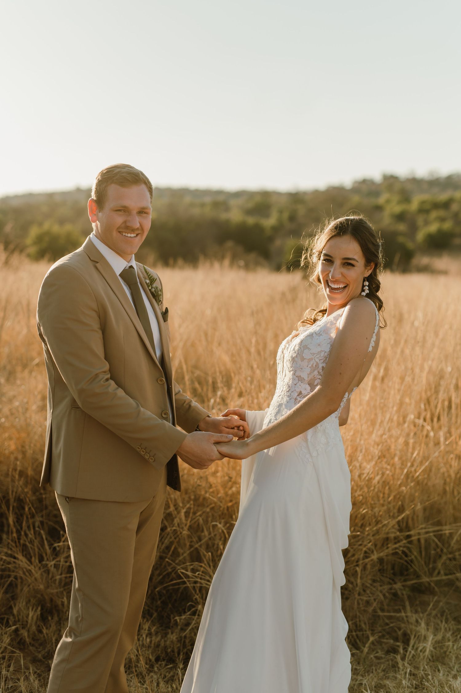 Newlyweds hold hands and smile at each other in a sun-lit field during golden hour.