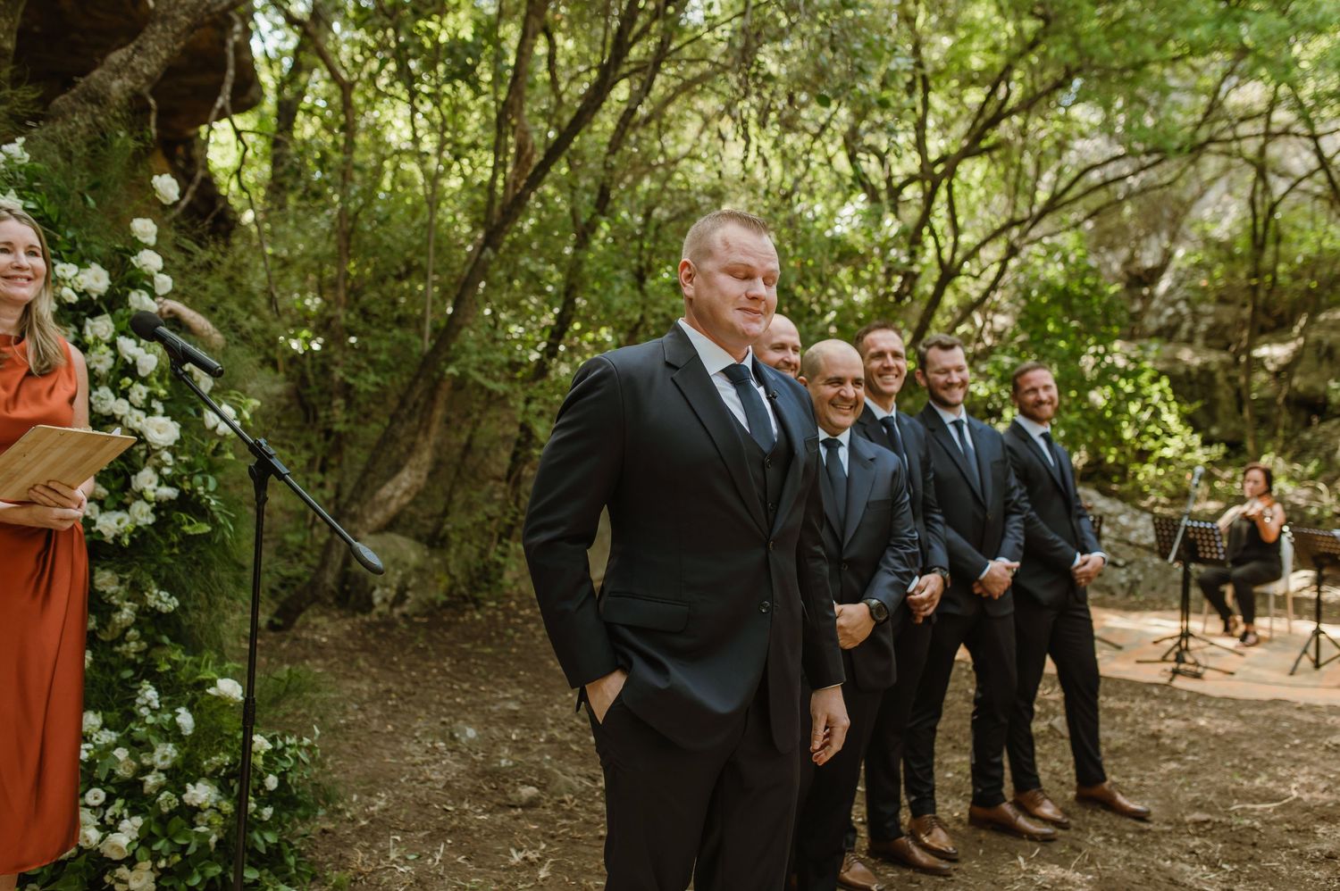 Wedding party members stand in formation along a tree-lined ceremony path.
